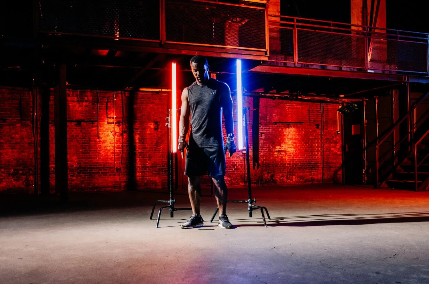 Een man in sportkleding in een industriel gebouw met rode en blauwe neonlichten, alsof hij zich voorbereidt op een workout of fotoshoot.