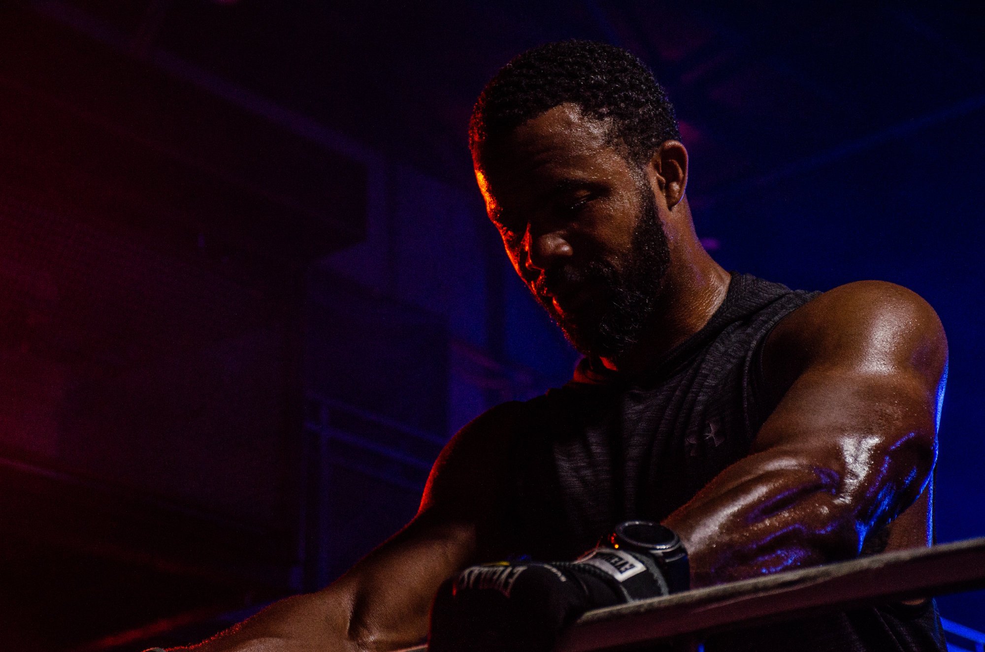 Mannelijke atleet die zweten en zich voorbereidt op een workout in een sportschool, gekleed in sportkleding en een horloge, met donkere achtergrond en gekleurde sportverlichting.