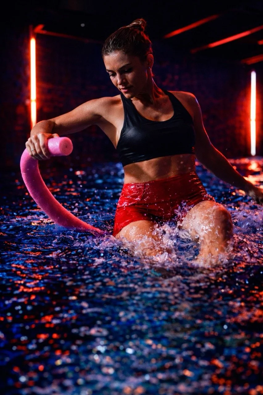 Vrouw in sportkleding die met een roze waterslang in een verlichte, neon-lichte zwembadomgeving oefent.