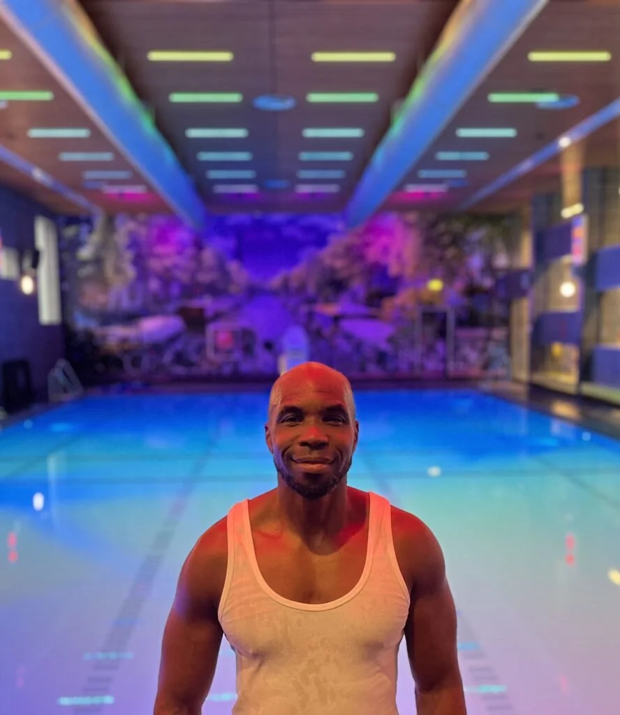 Een man in een wit tanktop staat voor het olympisch zwembad met kleurrijke verlichting en bloemen op de achtergrond.