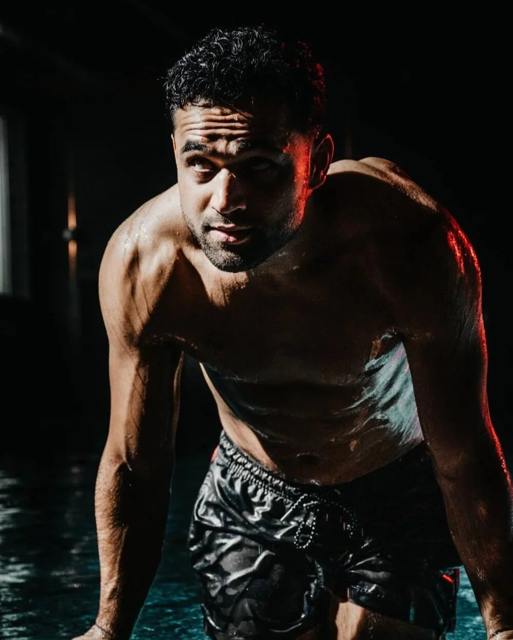 Een shirtloze man met natte huid, in een houding alsof hij klaar is om te sporten, set tegen een donkere achtergrond met rode en blauwe verlichting.