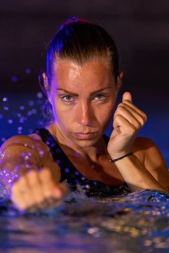 Vrouw in een zwembad die een nestelachtige houding aanneemt, met nat haar en waterdruppels op haar gezicht.