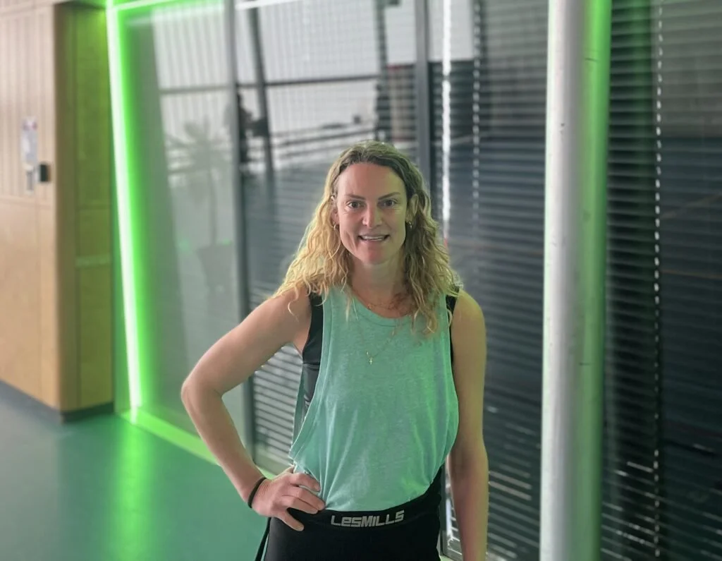 Vrouw met wapperend haar en sportkleding, poseert binnen in een modern gebouw met groene verlichting.