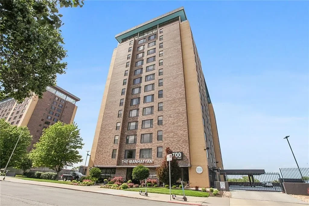 A tall beige and brown apartment building named The Manhattan, with several windows, trees, and a sidewalk in front.