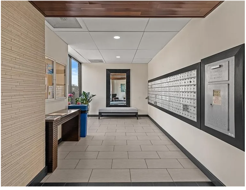 An indoor lobby area with a row of mailboxes on the right wall, a mirror and a bench at the end, a window on the left, and some decorative plants near the window.