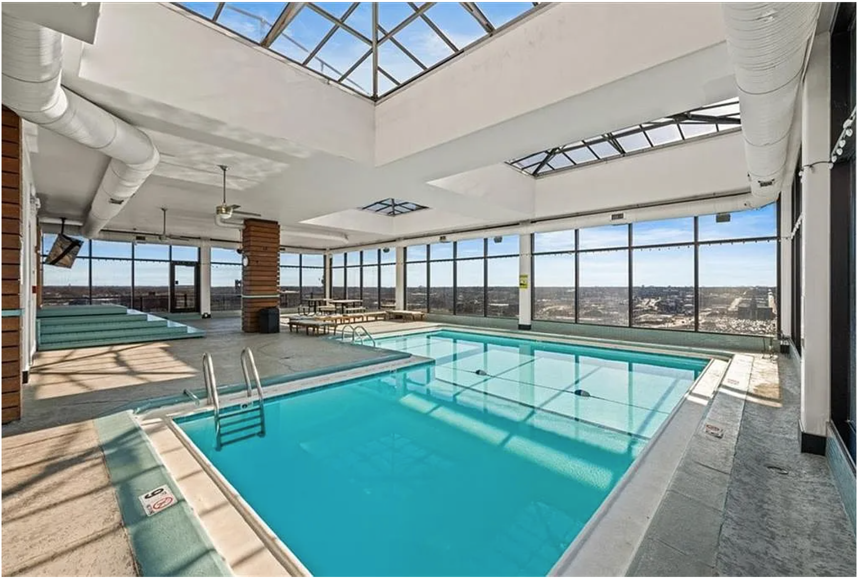 Indoor swimming pool area with large windows, skylights, and city view, including a hot tub, lounge chairs, and overhead ceiling fans.