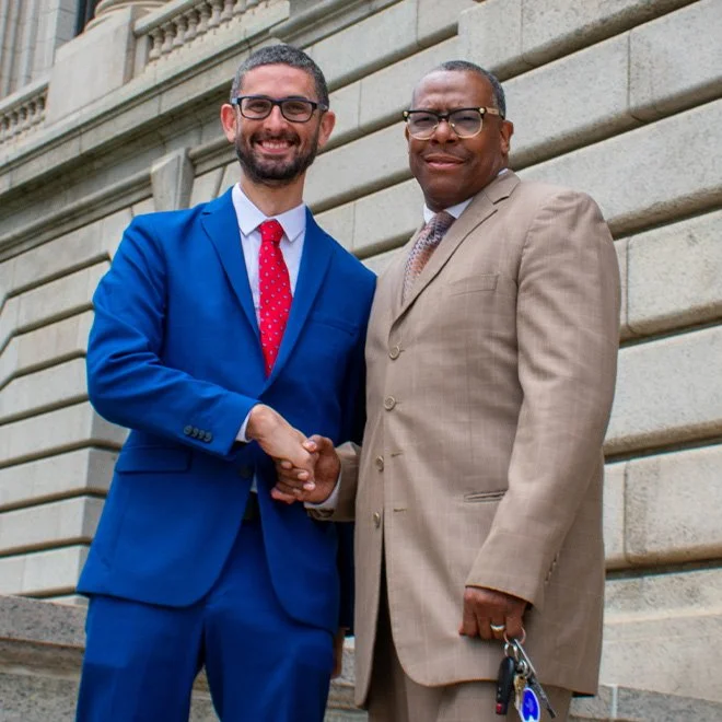 Two men in suits shaking hands and smiling outdoors in front of a stone building.