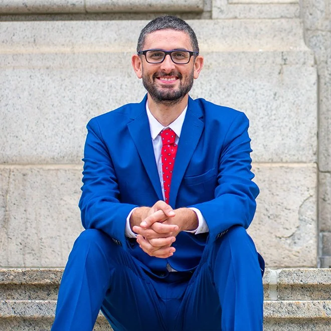 A man in a blue suit with glasses and a red polka dot tie, sitting on stone steps outside a building with stone walls, smiling and clasping his hands.