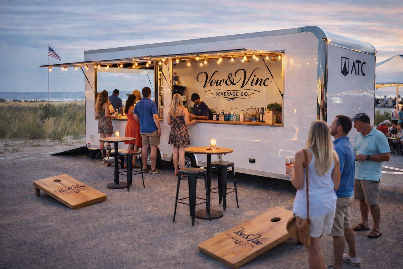 People standing at a mobile bar named 'Vow & Vine Beverage Co.' at sunset on a beach. The bar has string lights, with some patrons holding drinks. Two wooden cornhole boards are on the ground, and a tent is visible in the background.