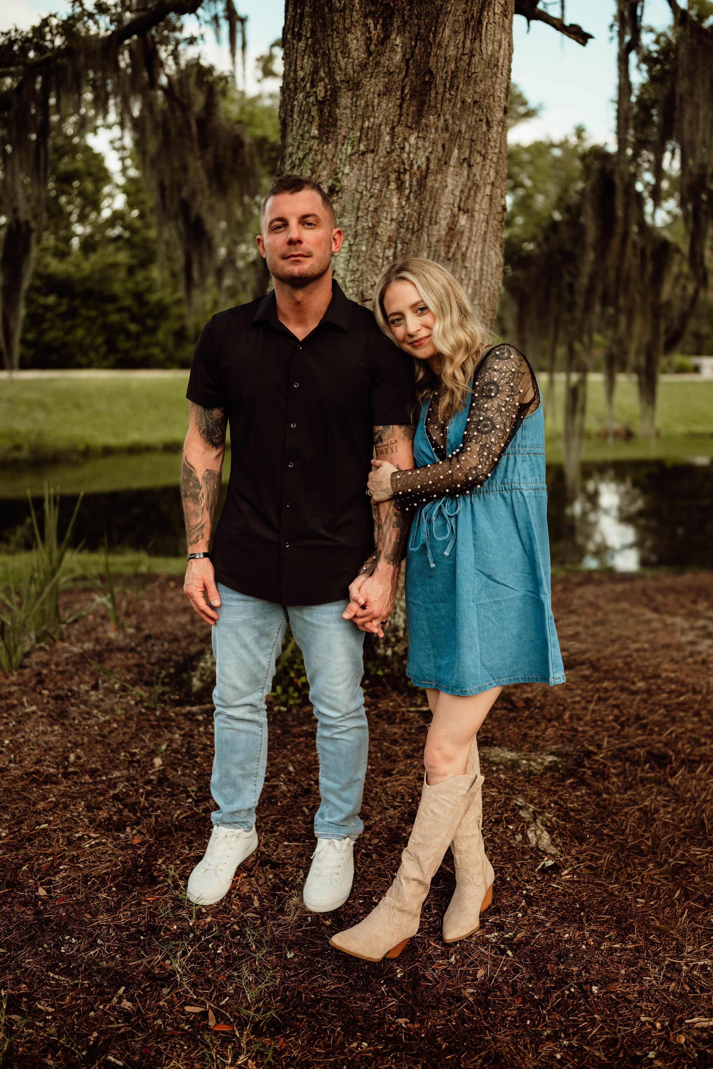 A young couple standing near a large tree by a pond, holding hands, in a park setting.