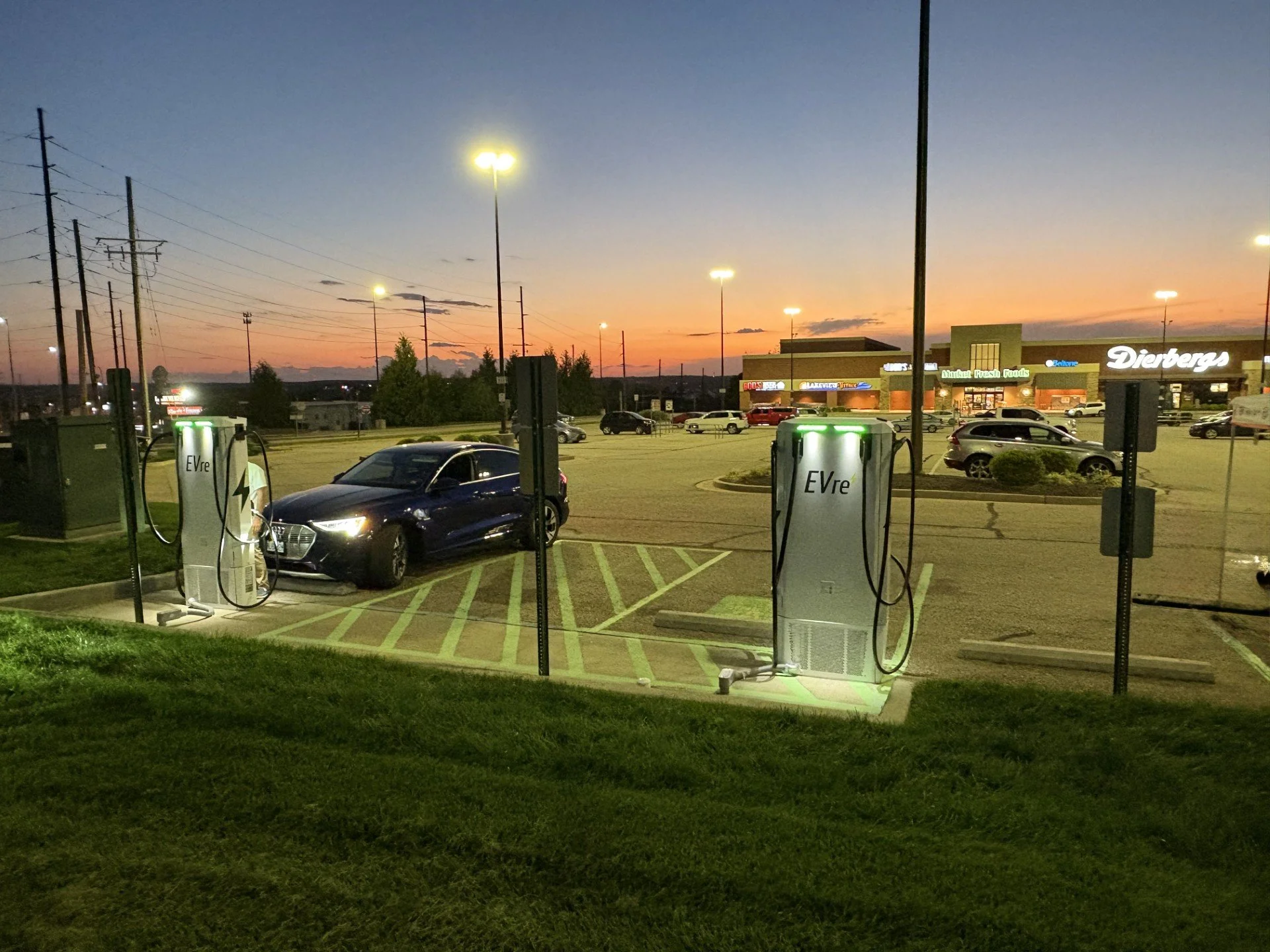 An electric vehicle charging station outside a shopping mall at sunset, with a dark sedan plugged into one charger and multiple cars in the parking lot.
