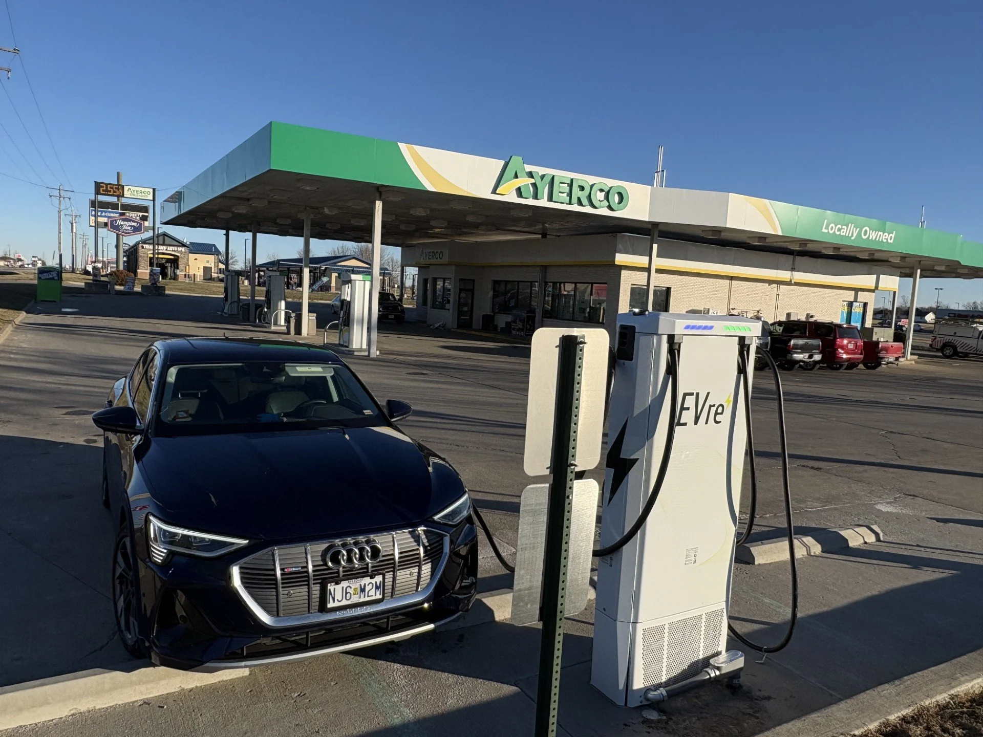 Black Audi electric vehicle charging at an Ayerco gas station with electric vehicle charger in the foreground and gas station building in the background.