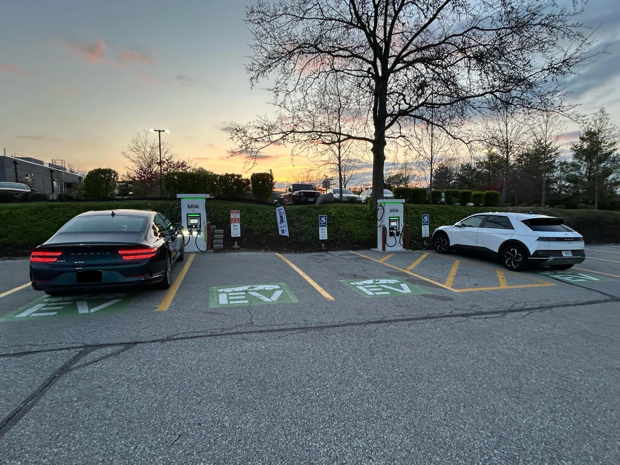 Two electric cars parked at charging stations in a parking lot during sunset, with trees and a sky with clouds in the background.
