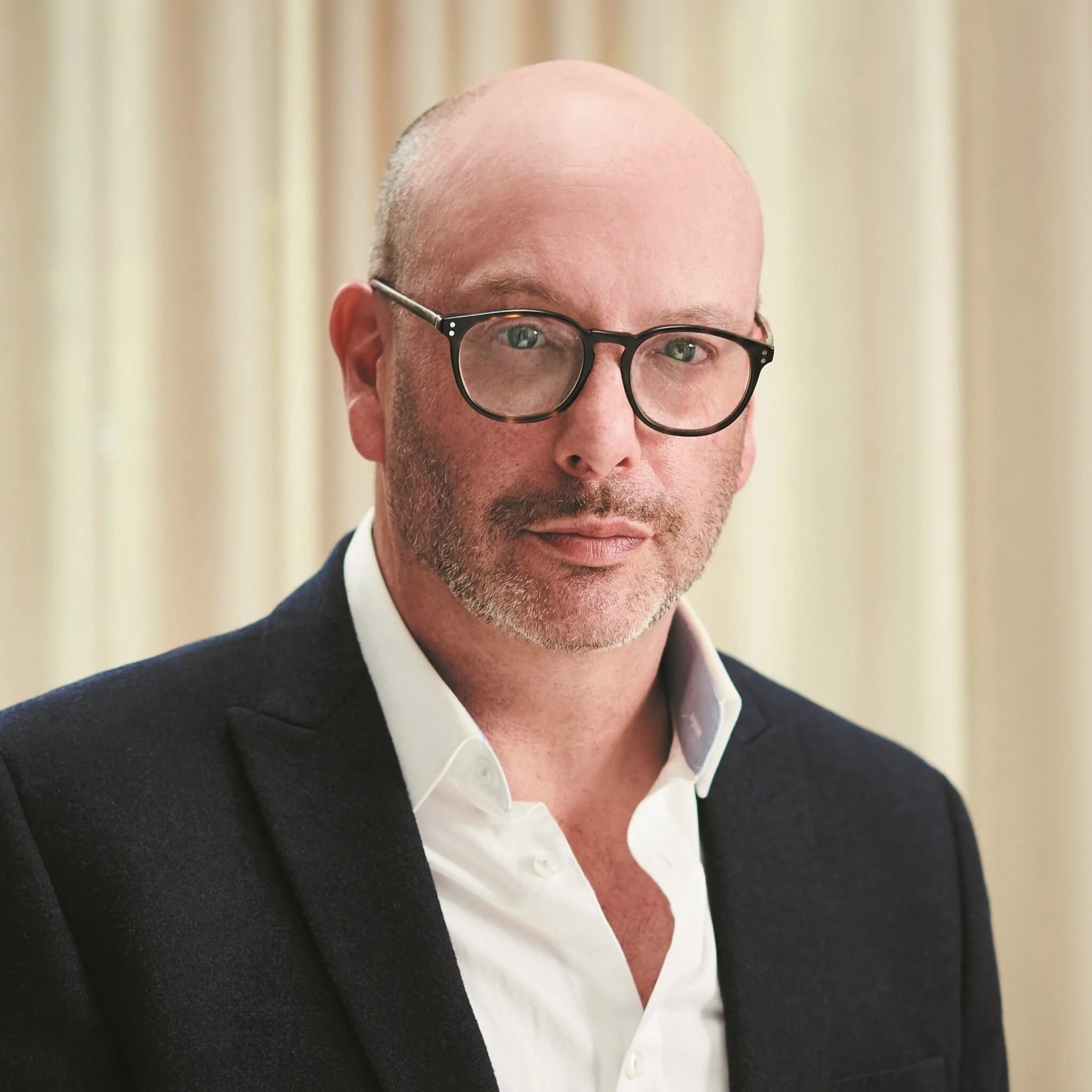 A middle-aged man with glasses, a beard, and a bald head, wearing a white shirt and a dark blazer, standing in front of beige curtains.