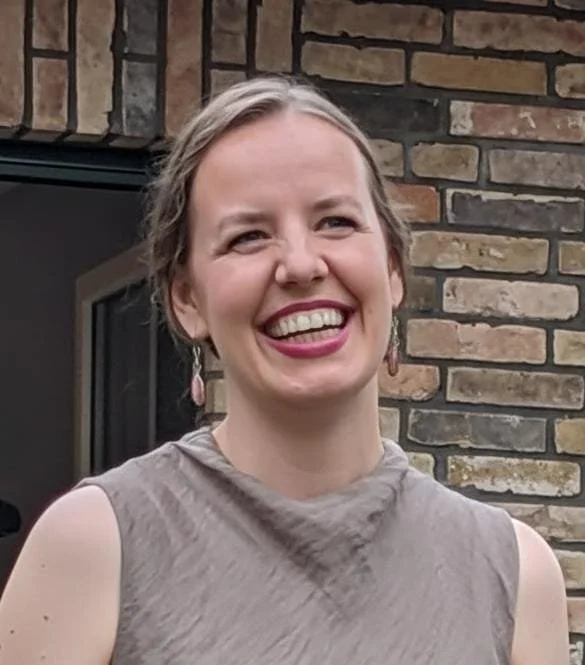 A smiling woman with light brown hair tied back, wearing a sleeveless beige top and pink earrings, standing outside in front of a brick wall.