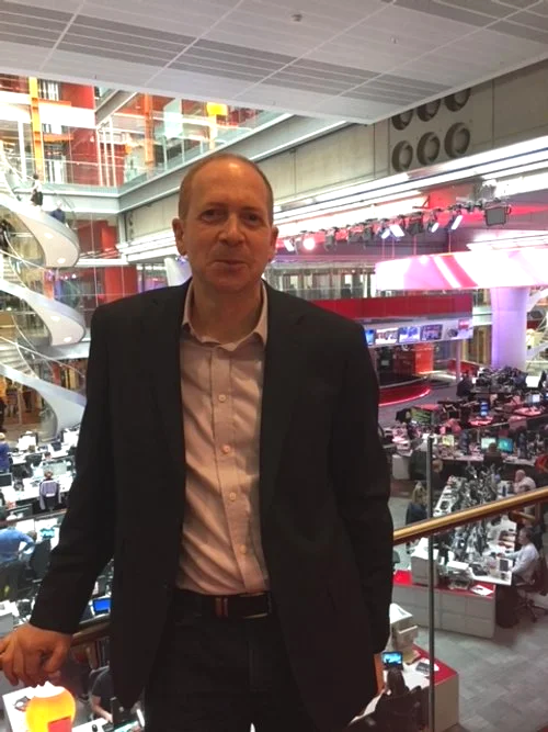 A man in a suit standing in a modern newsroom or media office with multiple computer workstations and large screens in the background.
