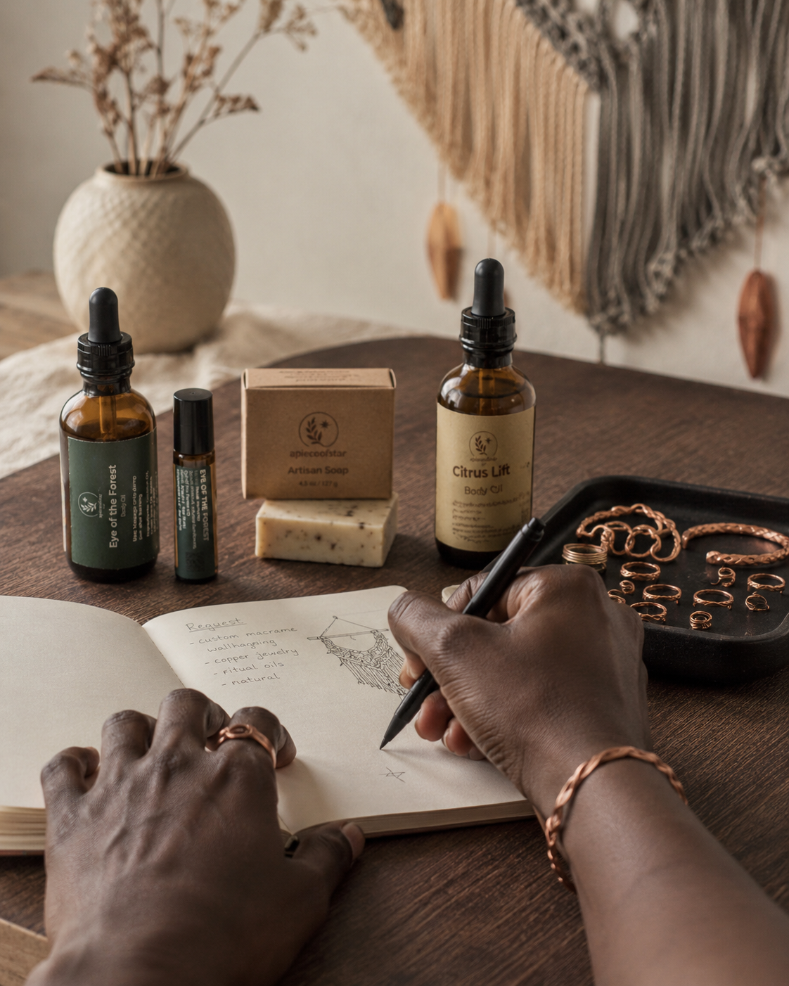 Hands writing in a notebook beside apieceofstar oils, a soap box, copper rings, and ritual tools.