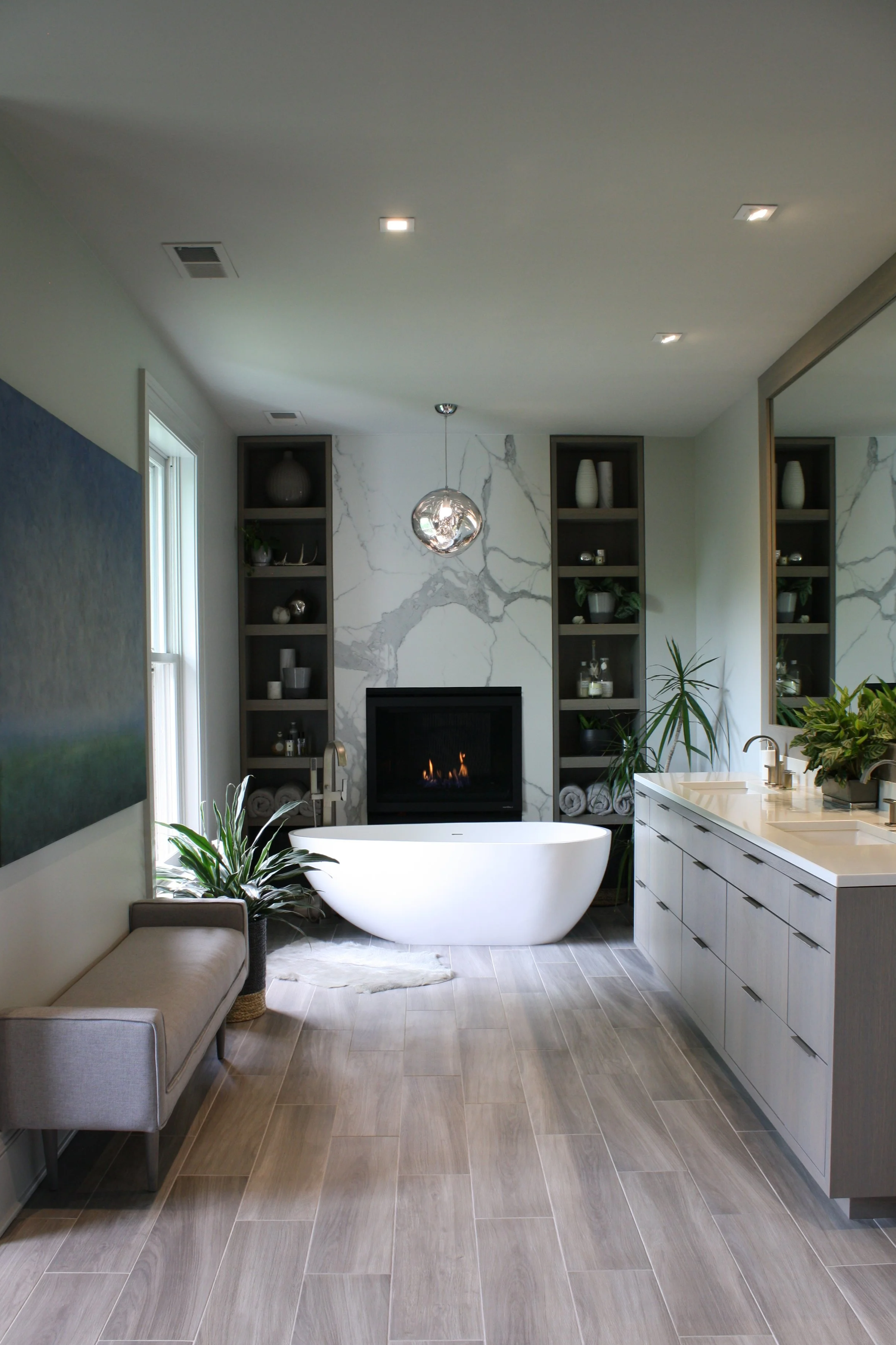 Modern bathroom with a white freestanding bathtub in front of a fireplace, marble accent wall, built-in shelves with decor, plants, large mirror, and vanity with sink.