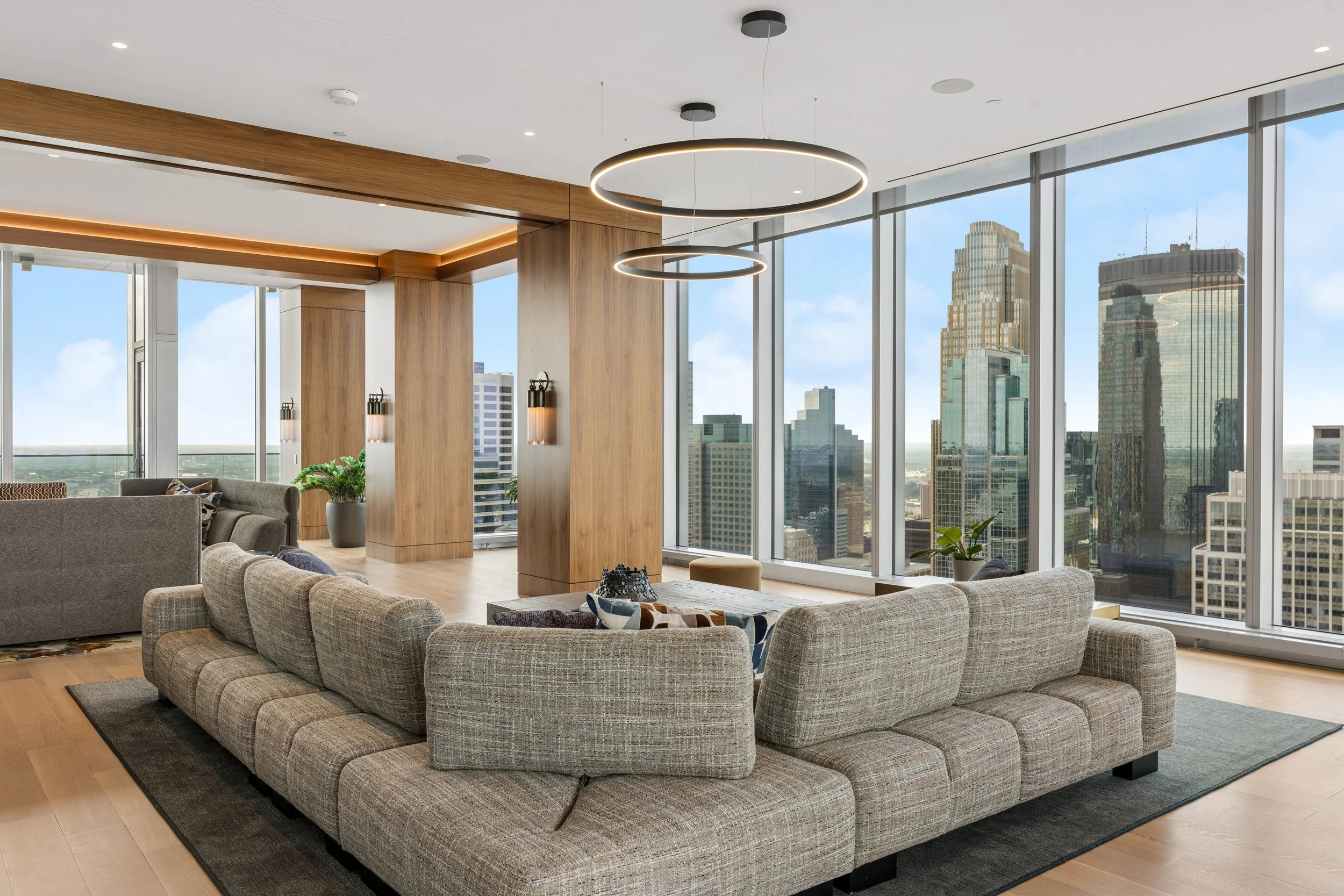 Modern living room with large glass windows showing a city skyline, beige sofas, a central wooden column, and circular pendant lights.