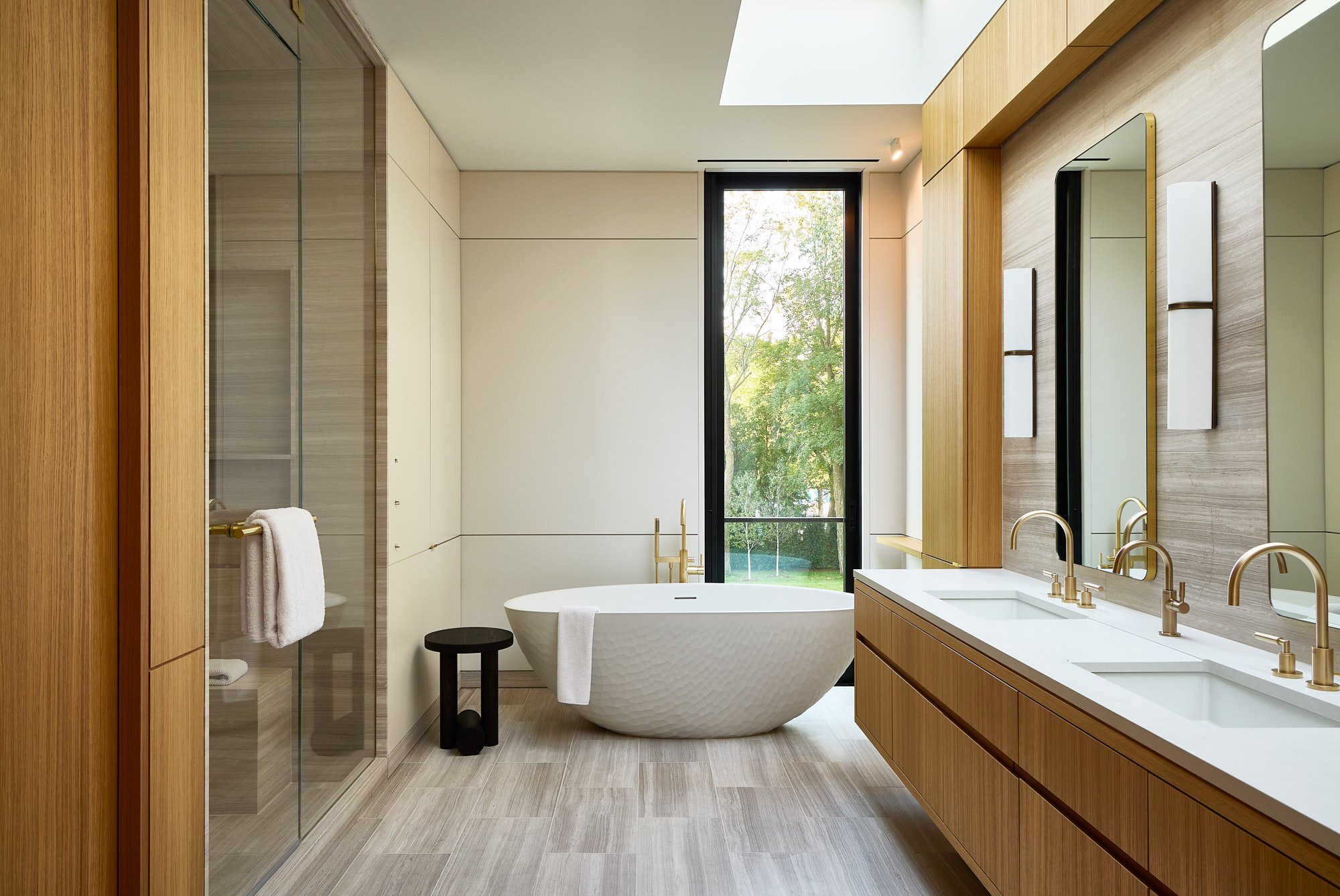 Modern bathroom with wooden accents, a freestanding bathtub, a double sink vanity, and large window showing green trees outside.