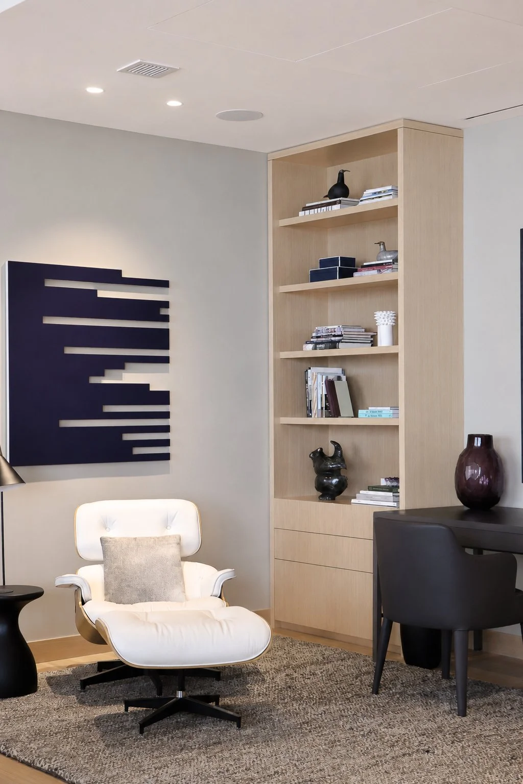 Modern living room corner with a white lounge chair, a black side table, a dark gray chair, and a tall wooden bookshelf with decorative items and books. A beige rug is on the floor, and a black wall art piece resembling piano keys is mounted on the l