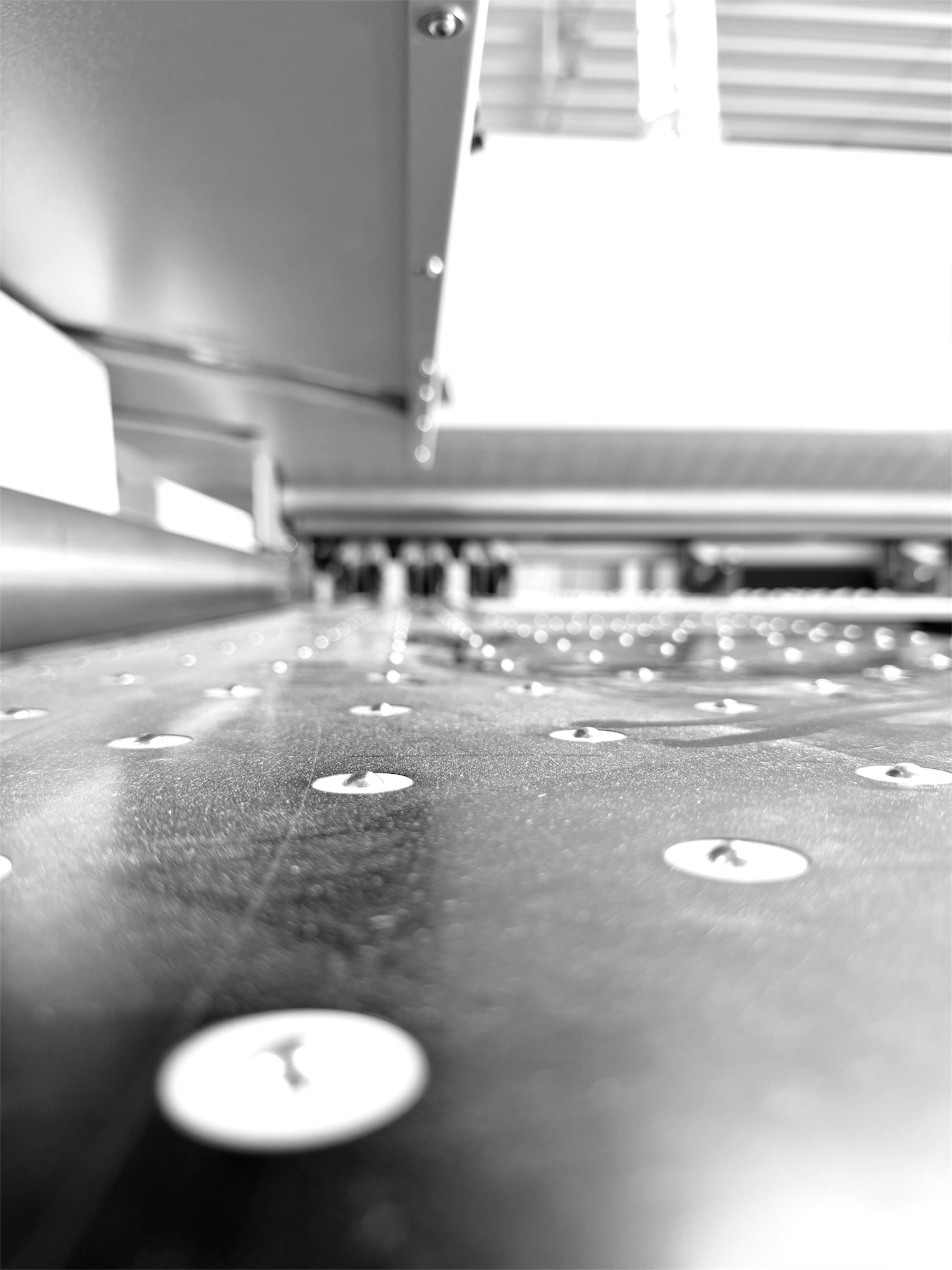 Close-up of a metal surface with evenly spaced rivets or bolts, with a part of a metallic structure above and a row of metal slats in the background.