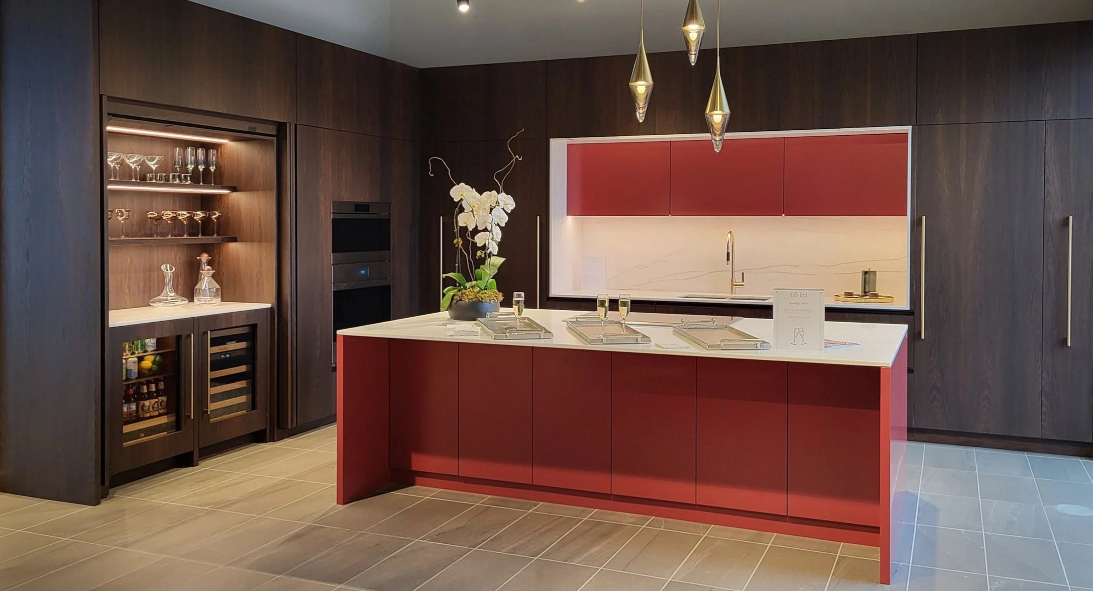 Modern kitchen with dark wood cabinets, a white marble island with red accents, a built-in wine fridge, and decorative glassware, chandelier, and floral arrangement.