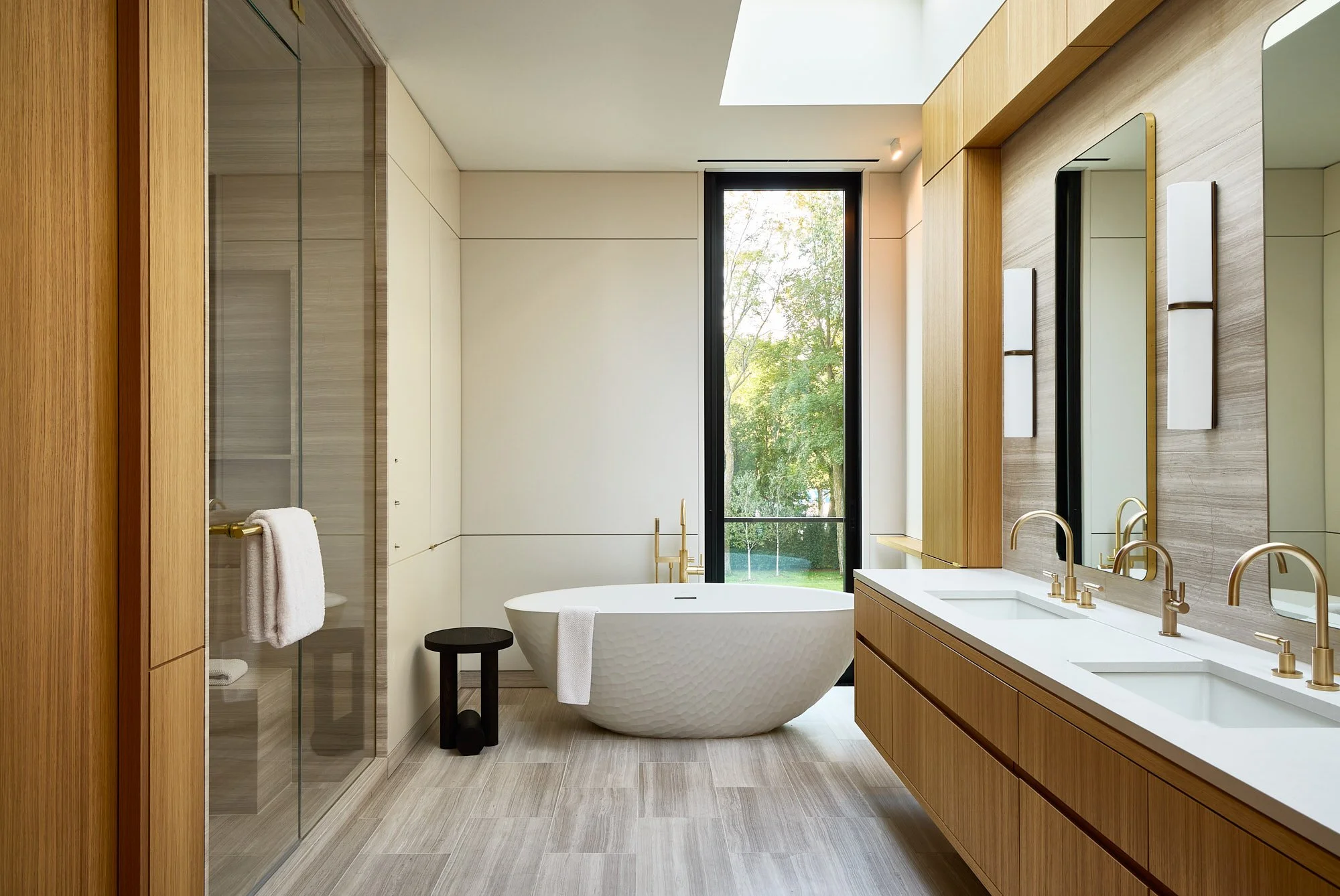 Modern bathroom with a freestanding bathtub, double vanity with two sinks, large mirror, natural wood cabinets, and a window with a view of greenery.