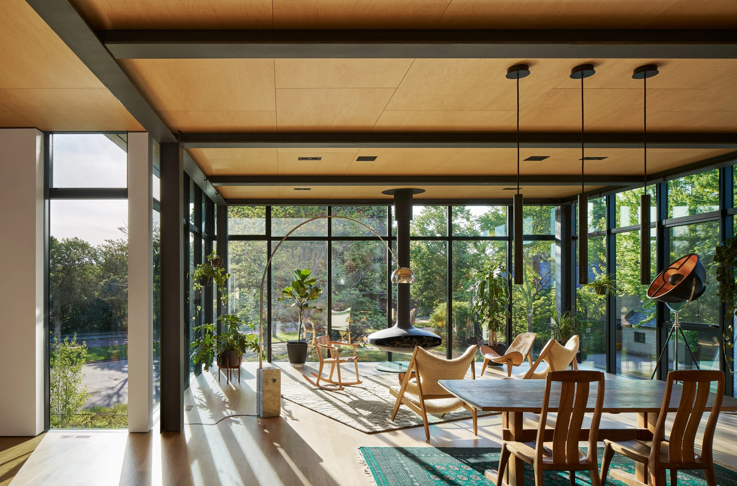 Interior of a modern home with floor-to-ceiling windows, wooden ceiling, and contemporary furniture, overlooking a green outdoor landscape.