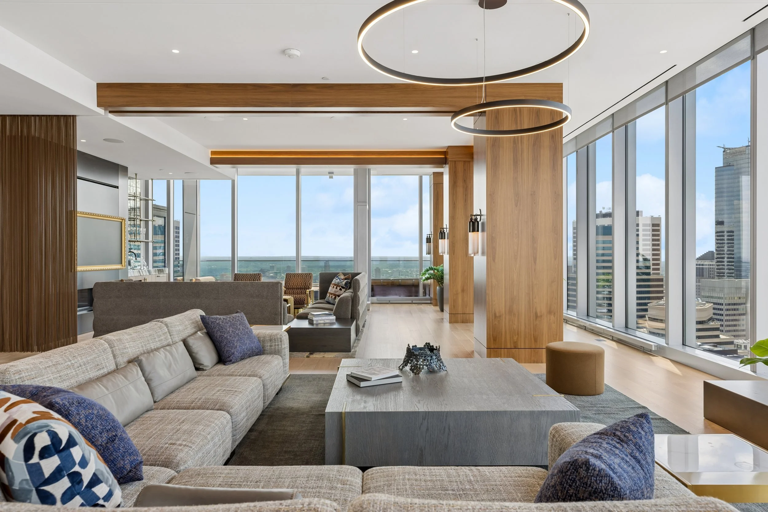 Modern living room with large windows, light wood floors, gray and beige sofas, and decorative pillows. There are two circular pendant lights hanging from the ceiling and a corner with a built-in bar area with glass shelves and a window view of high-
