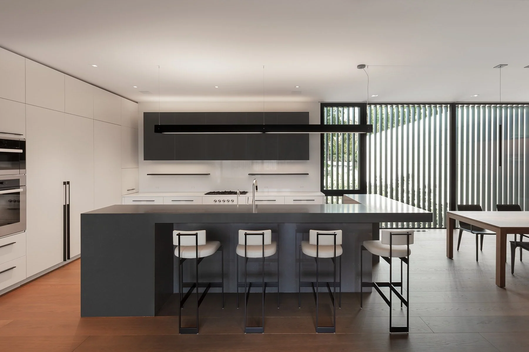Modern minimalist kitchen with white cabinets, a black island with white bar stools, and large windows with vertical blinds.