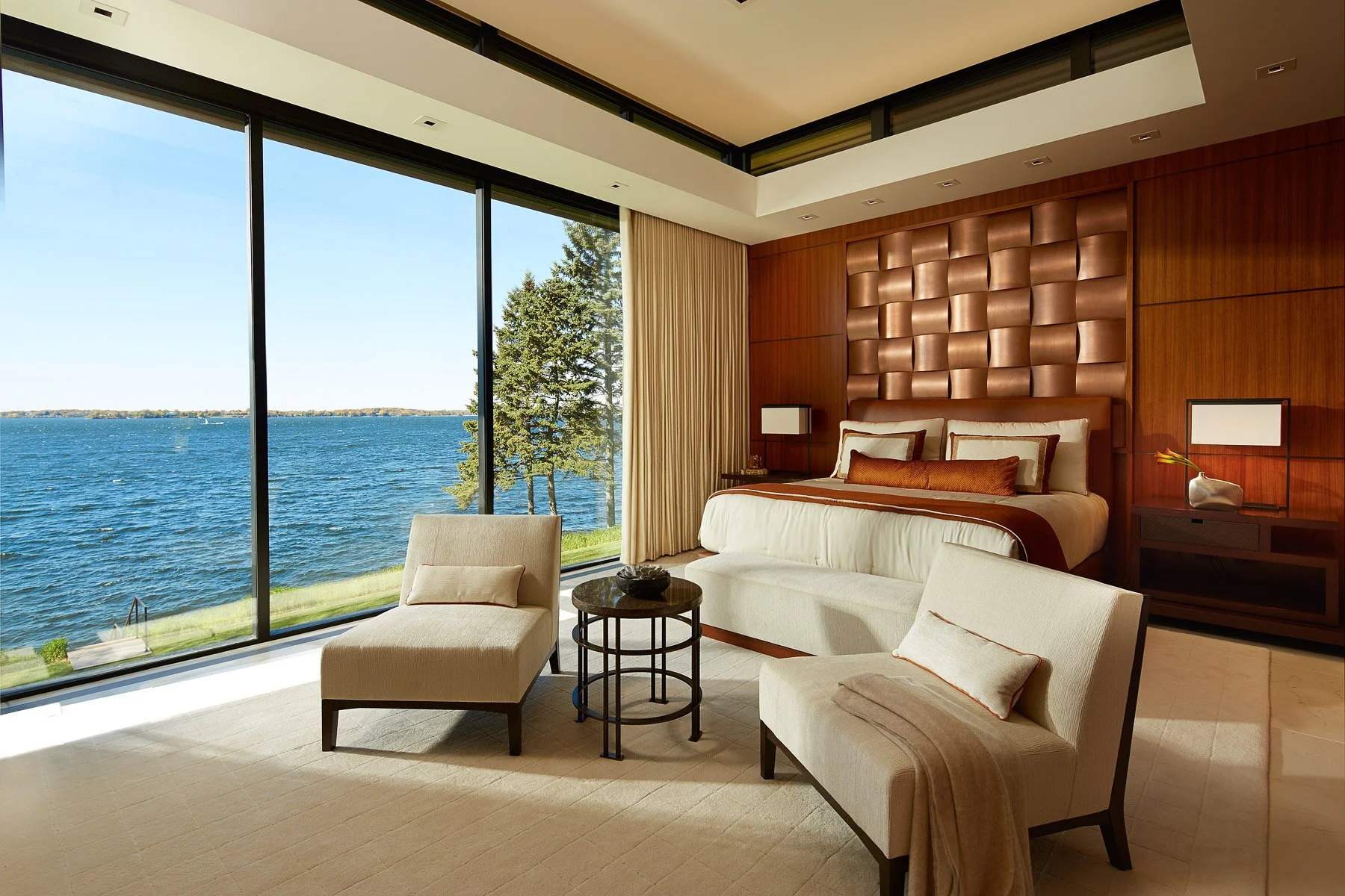A luxurious bedroom with a large window overlooking a body of water and trees outside. The room features a bed with a padded headboard, side tables, lamps, and comfortable chairs and a small round table near the window, all with a modern and elegant 