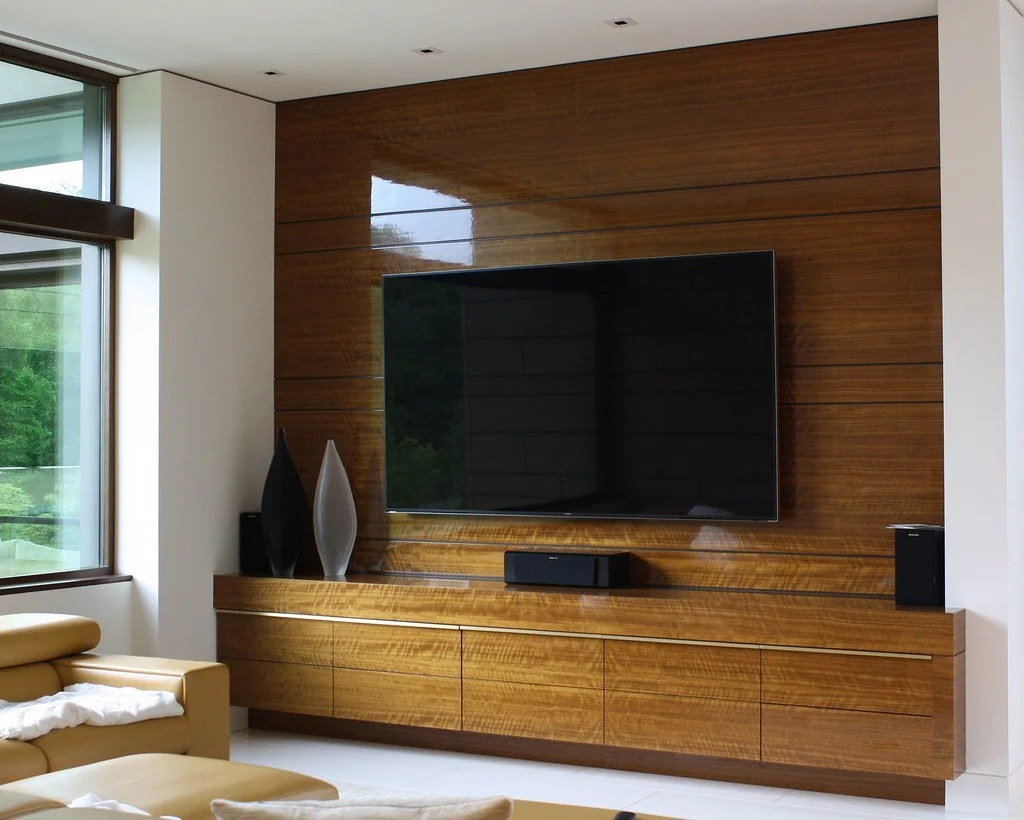 Modern living room with a large flat-screen TV mounted on a wood-paneled wall, decorated with vases, next to a window with a view of greenery, and a beige sofa with white cushions.