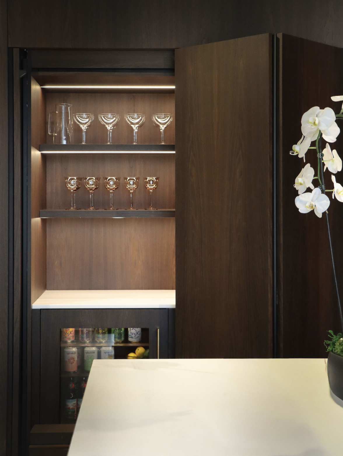 A wooden bar cabinet with glasses on shelves and canned beverages in a mini fridge below, next to a white countertop and a potted white orchid plant.