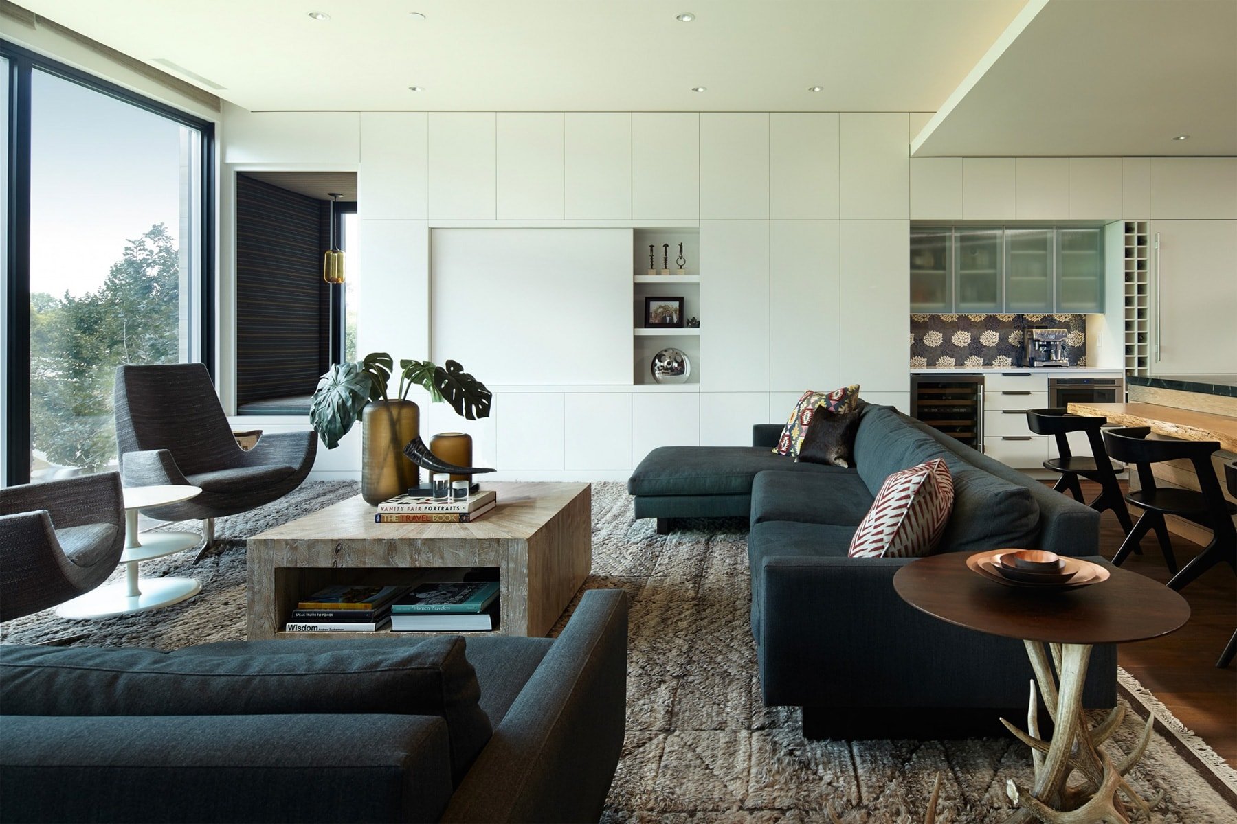 Modern living room with a fireplace, a flat-screen TV, a wooden and stone wall, and large windows with a view of trees.