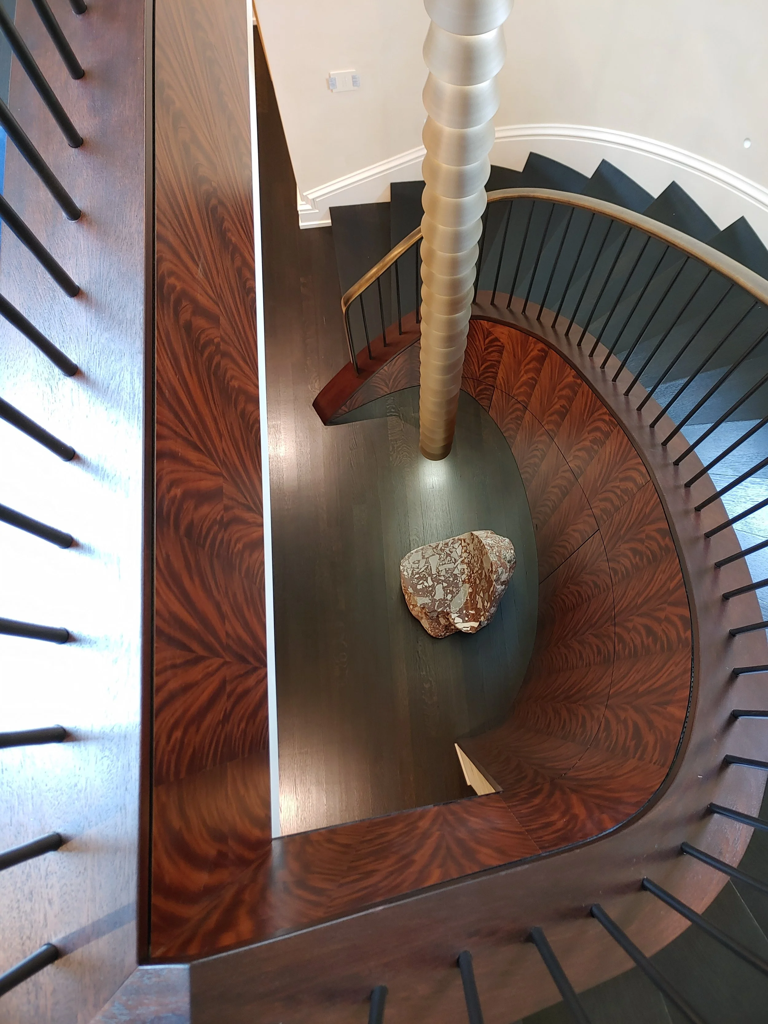 Spiral staircase viewed from above, with wooden railing and black steps, next to a stone sculpture and a green floor.