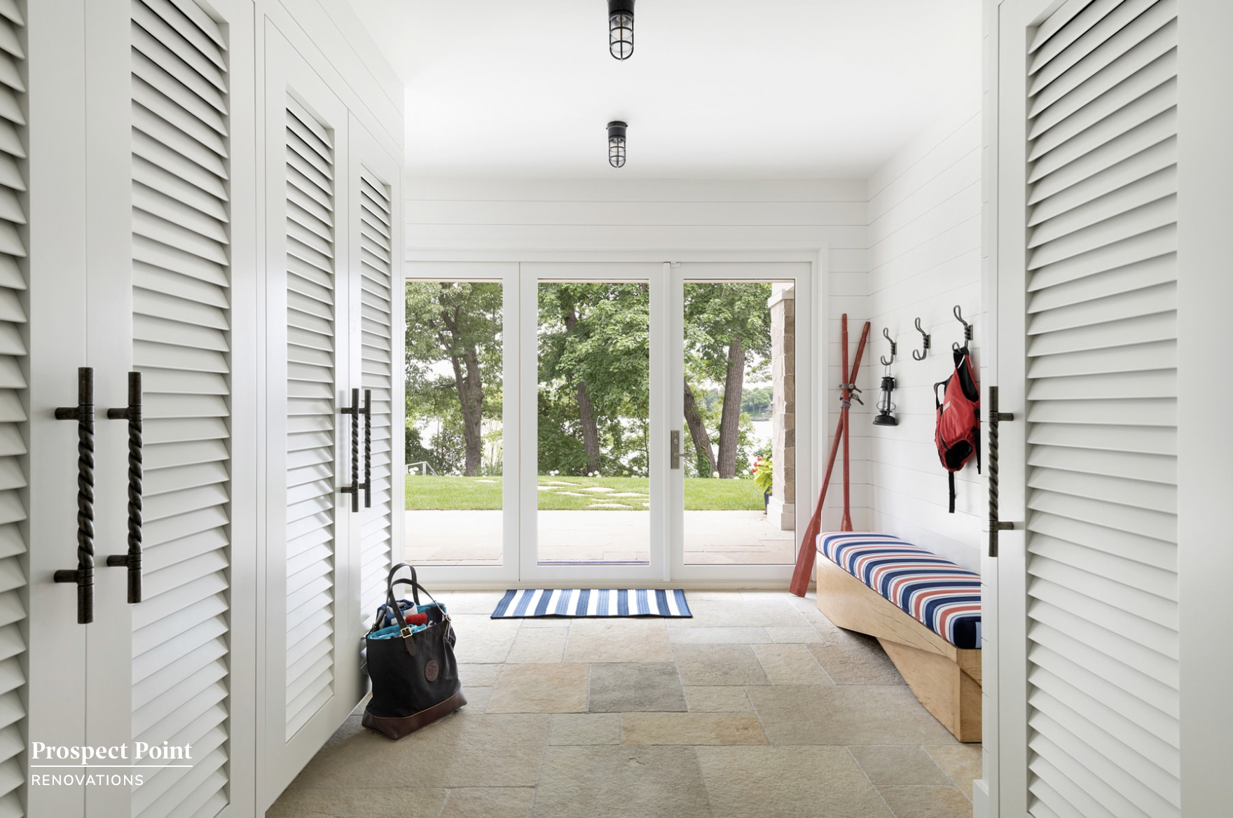 Entryway with white louvered closet doors on both sides, a striped cushioned bench, a black tote with items, and outdoor scenery visible through glass sliding doors.