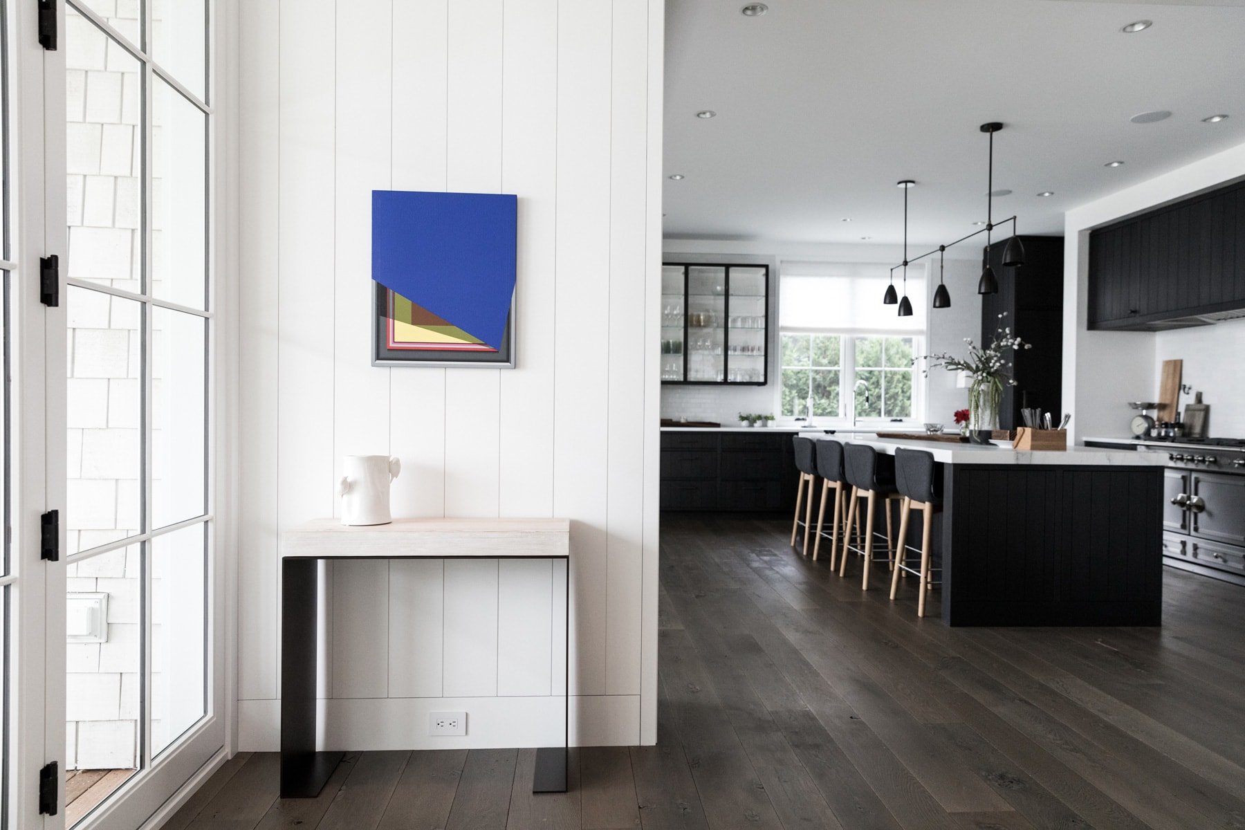 Modern kitchen with black cabinets, white countertops, and a kitchen island with four bar stools. Large windows allowing natural light. Decorative branches in a vase on the kitchen island. Minimalist lighting fixtures hanging from the ceiling.