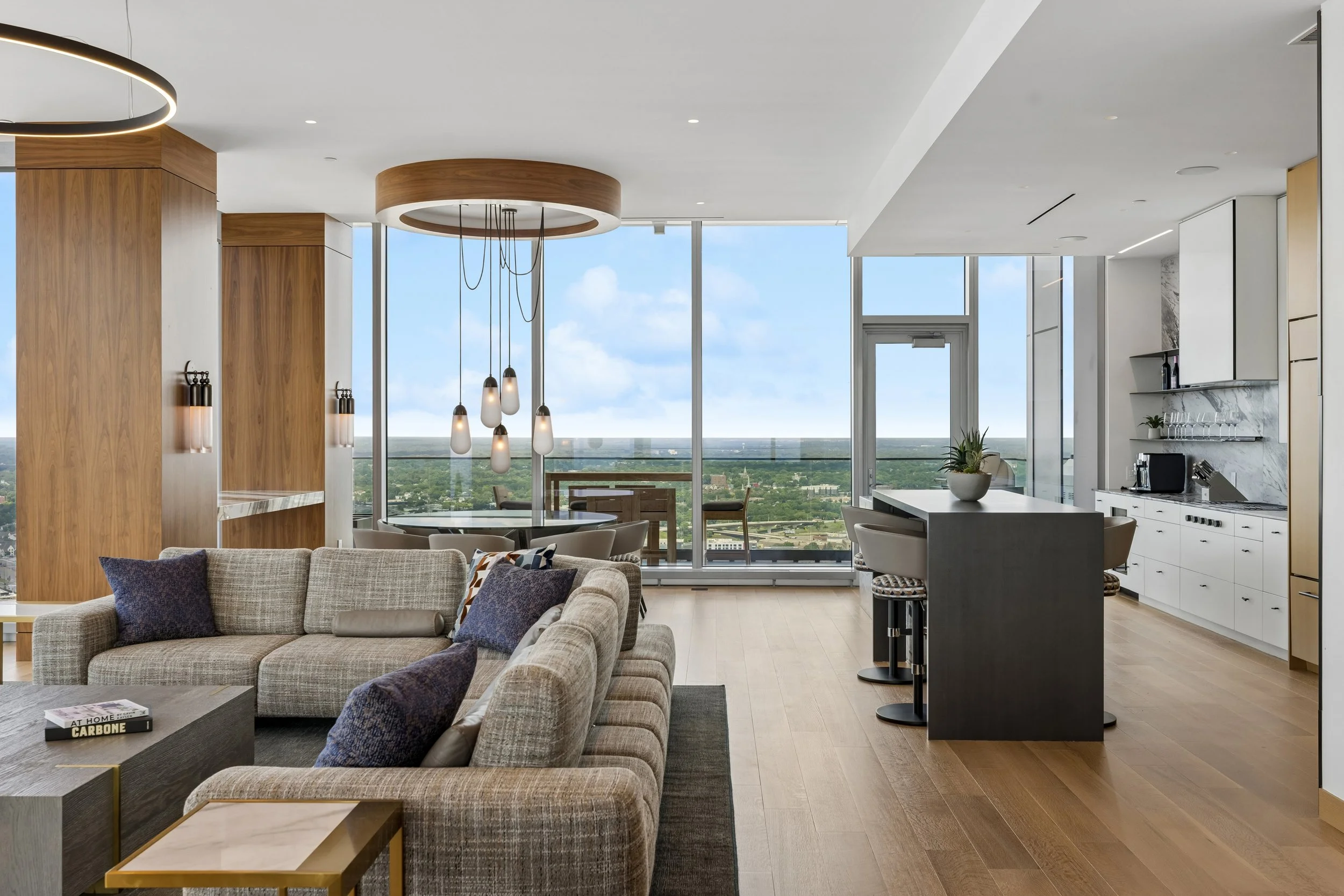 Modern living room and kitchen with large windows showing cityscape and blue sky, beige sectional sofa, coffee table, pendant lights, kitchen island with barstools, and wooden cabinetry.