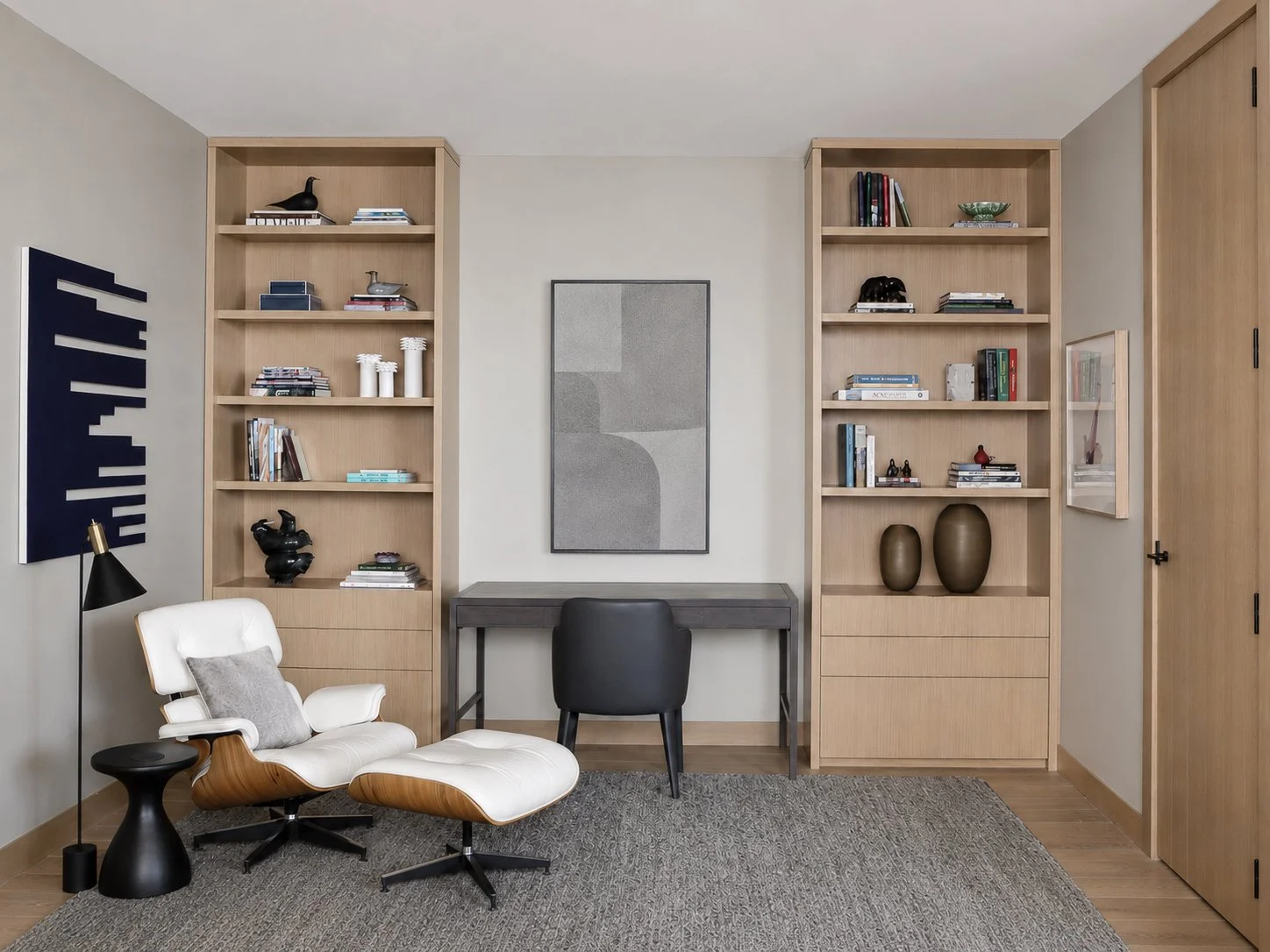 Modern home office with wooden bookshelves, a black desk, a white lounge chair with a gray pillow, a black floor lamp, and decorative vases and sculptures.