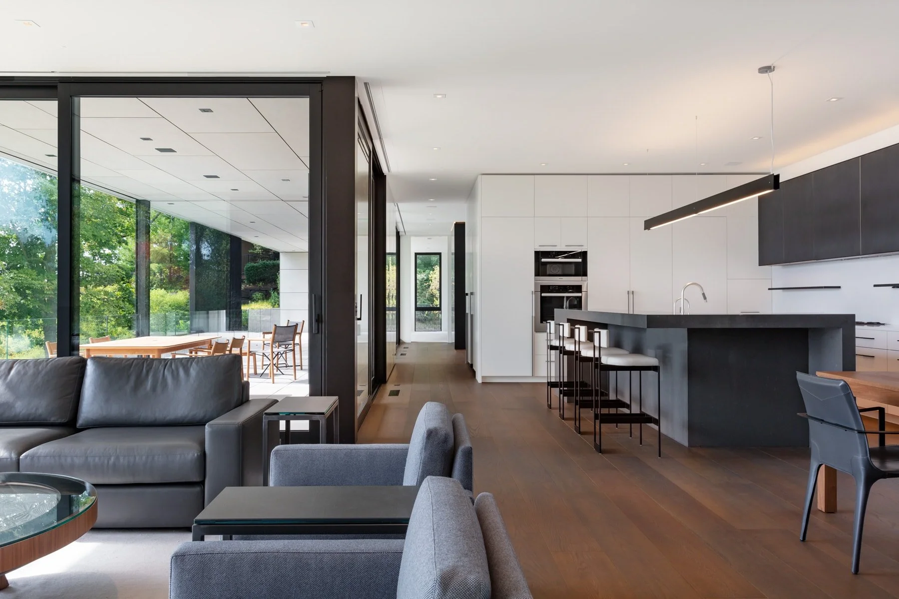 Modern open-concept kitchen and living room with large windows, gray and black furniture, wooden flooring, and a balcony with outdoor dining.