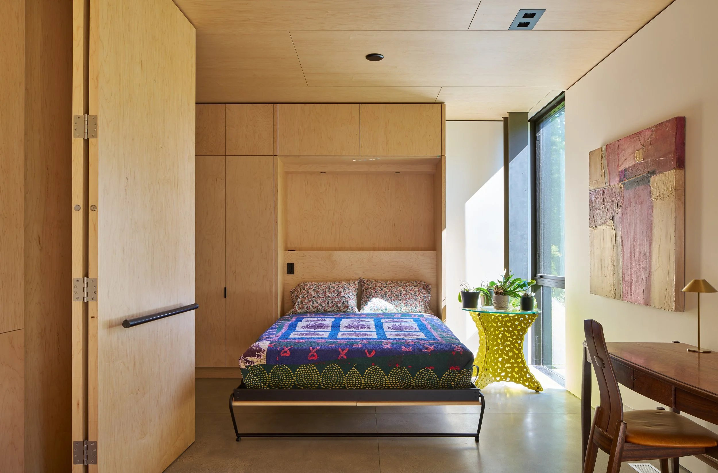 Interior of a modern bedroom with light wood paneling, a bed with colorful quilt and floral pillows, a yellow side table with potted plants, a wooden desk with a chair, and abstract artwork on the wall by large windows.