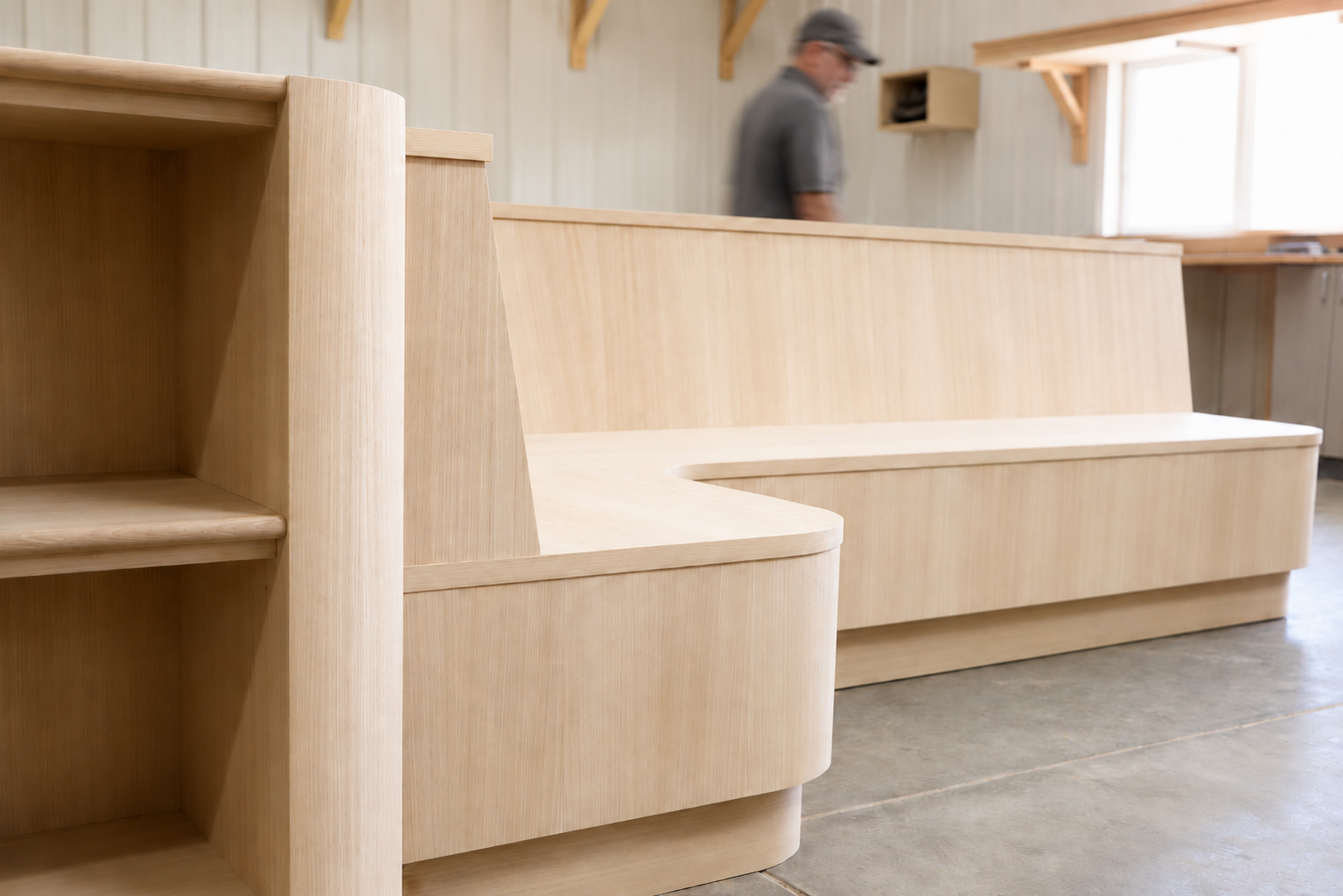 Unfinished light wood furniture, including shelves and a bench, in a workshop with a man in the background.