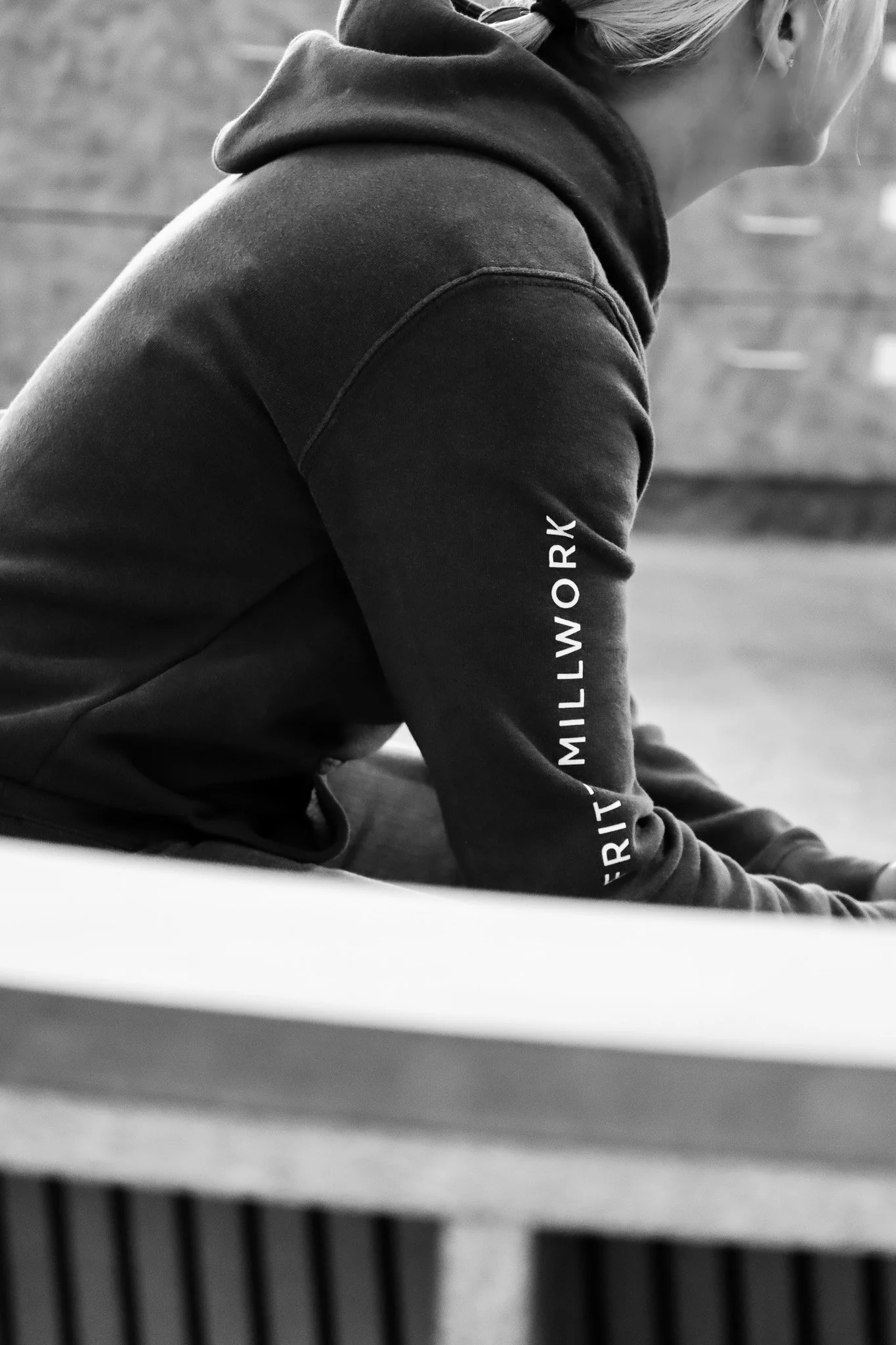 A person wearing a dark hoodie with the words Fritz Millwork printed on the sleeve, sitting at a table with a wooden surface, viewed from side, in a black and white photo.