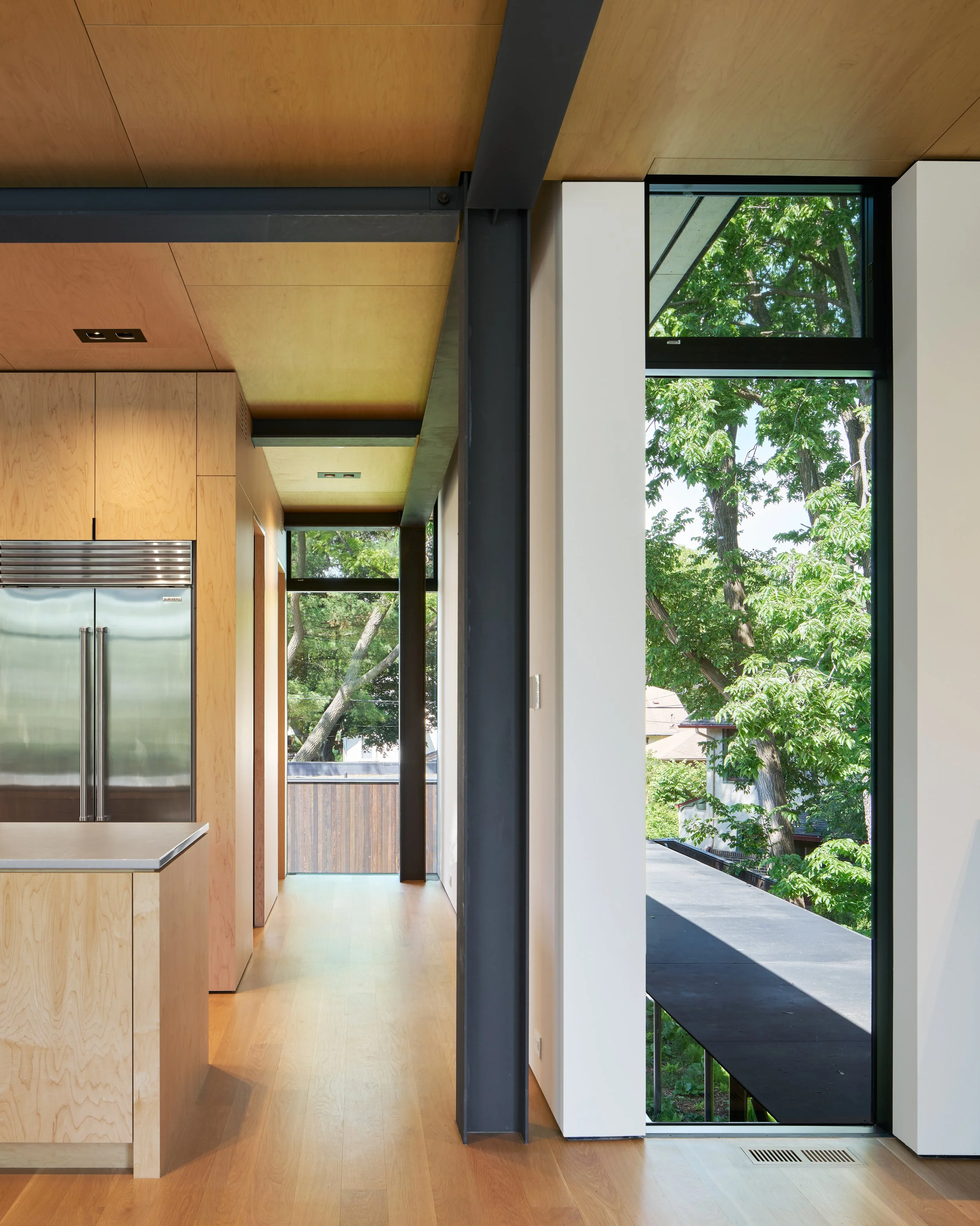 Modern interior with wooden cabinetry, hardwood flooring, large windows showing green trees outside, and black metal support beams.