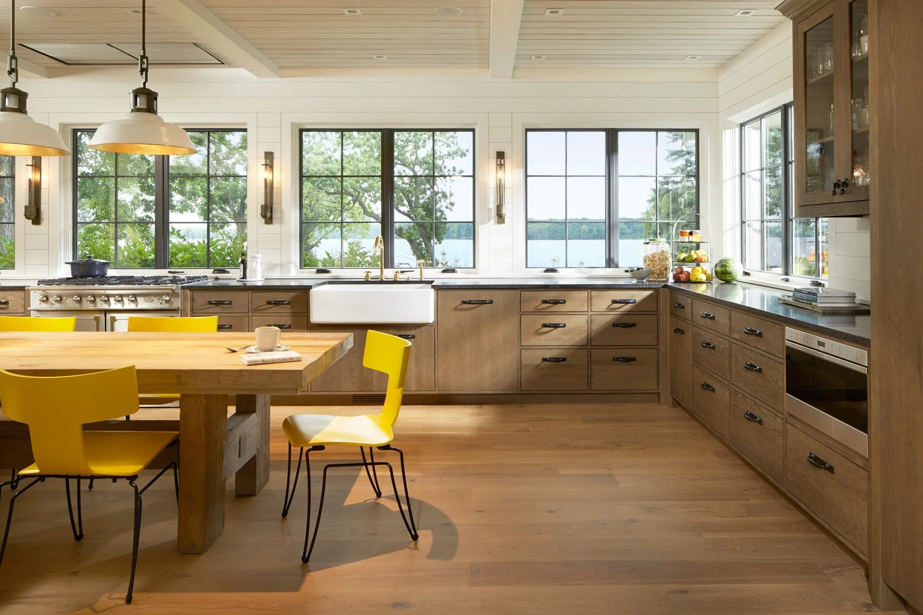 Modern kitchen with large windows overlooking a lake, wooden cabinets, a farmhouse sink, a wooden dining table with yellow chairs, and various kitchen items including fruits, a cookbook, and jars.