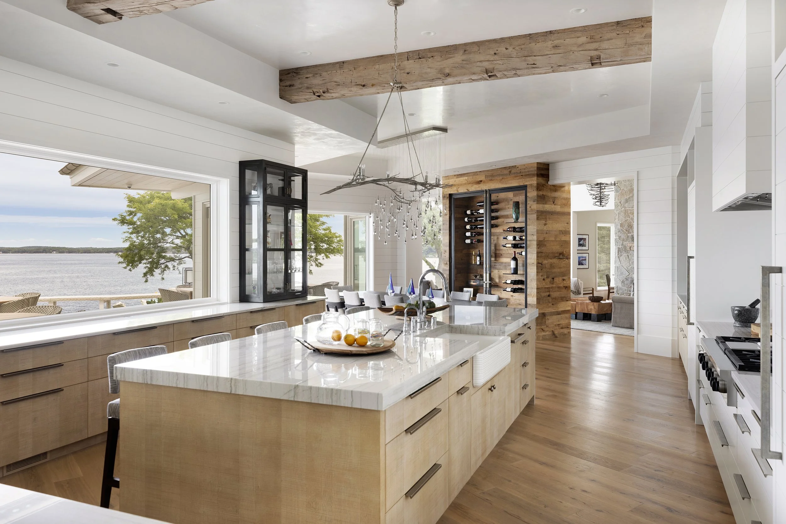 Bright kitchen with large island, wooden cabinets, wine cooler, and large window overlooking water and trees.