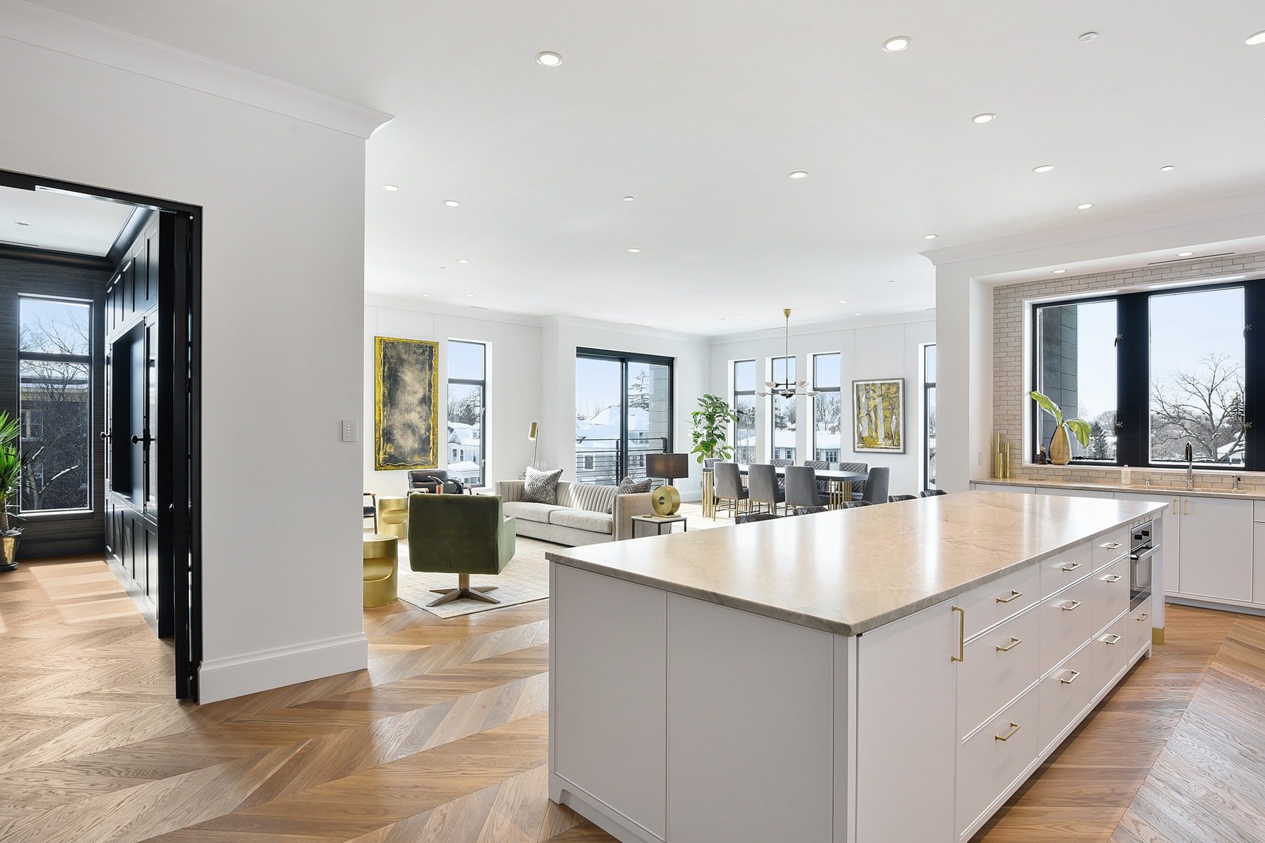 Open-concept living and dining area with white walls, large windows, and modern furniture, including a sofa, armchair, dining table with chairs, and a kitchen island with marble countertop, hardwood floors, and decorative artwork.
