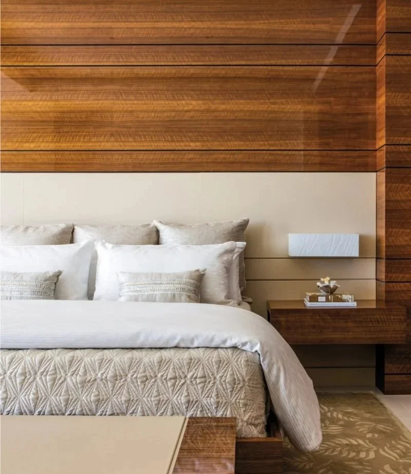 A nicely made bed with white and beige pillows, a beige quilted bedspread, in a room with wooden wall paneling and a white rectangular wall-mounted light fixture. A bedside table with decorative items is on the right side.
