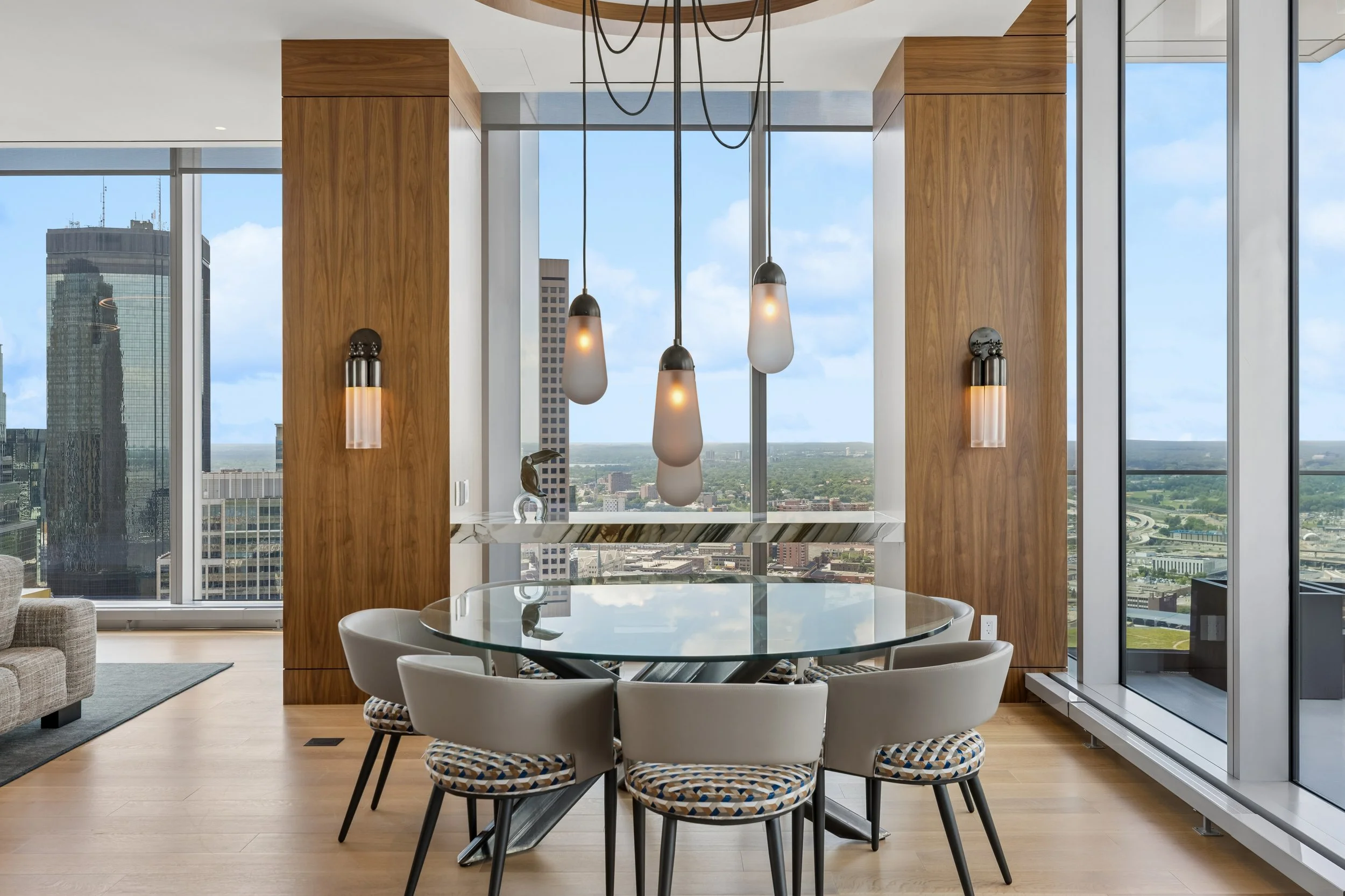 Modern dining area in a high-rise apartment with a round glass table, white chairs with patterned cushions, wooden wall panels, hanging pendant lights, large windows with city views and a blue sky.